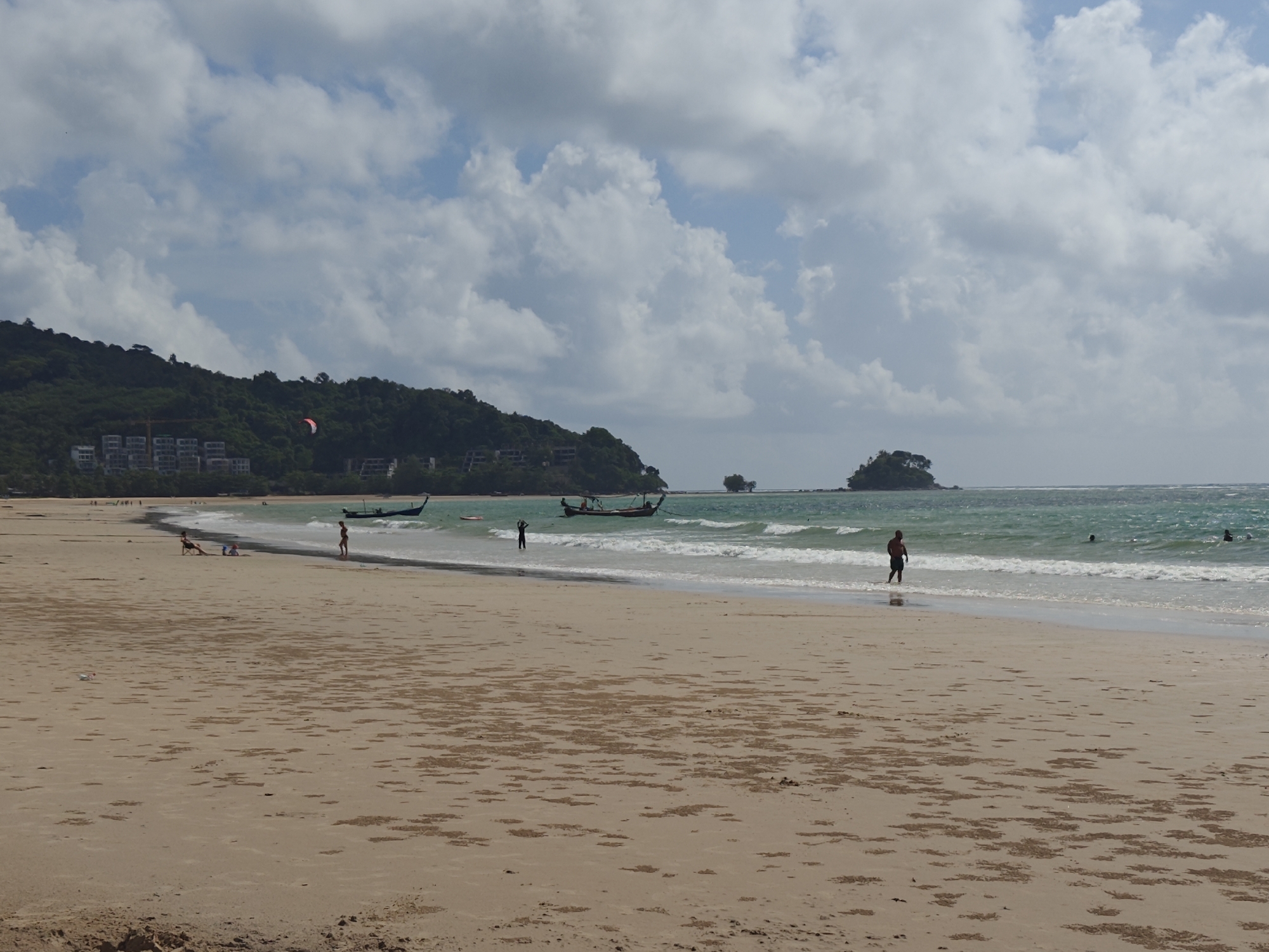 This is a wide shot of a sandy beach in Sakhu, Thailand, with the ocean and a tree-covered hillside in the background. The sky is filled with white, fluffy clouds. On the beach, several individuals are visible. In the foreground, the sand is dry and marked with small, scattered clumps. Further down the beach, near the water's edge, two people are standing, and two more are sitting. In the middle ground, a long-tail boat is anchored in the turquoise water, and another is further out. There are also several people in the water, swimming. The hillside in the background is densely covered with green trees, and several buildings are nestled amongst them. A kite is visible in the sky above the hillside. The scene depicts a typical day at a tropical beach.