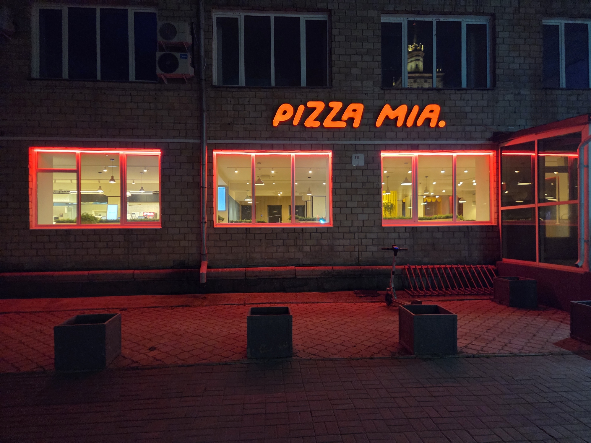 The image captures the exterior of a "PIZZA MIA." establishment in Chelyabinsk, Russia, at night. The building features a brick facade with several large windows illuminated by red neon lights outlining their frames. Above these, a larger set of windows on the second floor are dark, with one reflecting a distant tower. The "PIZZA MIA." sign is prominent, rendered in glowing orange lettering. In the foreground, a red-tiled walkway is visible, with three dark rectangular planters placed along it. To the right of the planters, a bicycle rack and a parked electric scooter are present, also bathed in the red light emanating from the building. The overall scene suggests a commercial area after closing hours, with the red lighting creating a distinct atmosphere.