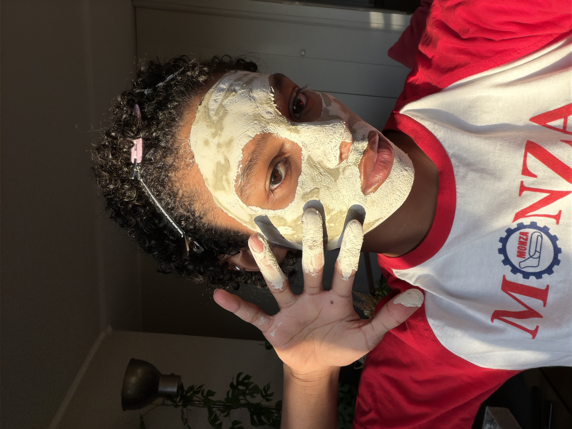 A young Black individual with curly dark hair, partially secured with silver and pink hair clips, is shown from the chest up. They are wearing a white raglan t-shirt with red sleeves and a red graphic on the front featuring the word "MONZA" and a circular logo. The individual has a pale, clay-based face mask applied to their face, covering their forehead, cheeks, nose, and chin. Their eyes are visible, with the left eye looking directly at the camera and the right eye slightly to the side. Their lips are also visible, appearing slightly glossy. The individual's left hand is raised, with fingers spread, and is also covered in the same white clay mask. The background appears to be an indoor setting, with natural light casting shadows. To the left, a portion of a plant and a dark-colored decorative object are visible. The overall lighting suggests it is either early morning or late afternoon. The image does not provide any information that would indicate the location in the city of Champs-sur-Marne, France.