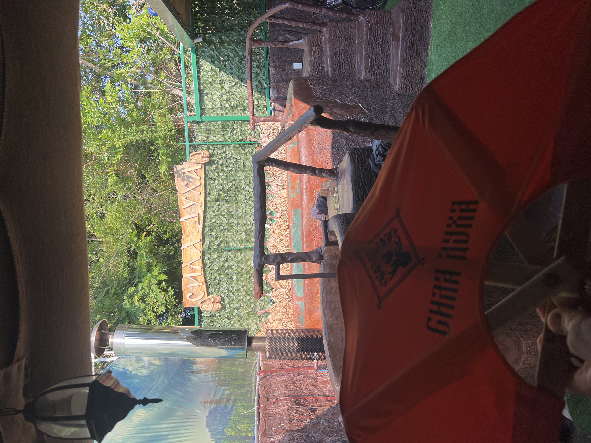 The image shows a rustic outdoor setting, possibly a patio or garden area, in what appears to be the Bac Natural Heritage Area Trang, Vietnam. The scene is somewhat obscured by an orange object, possibly a tent or canopy, with Cyrillic text and a graphic of a bear. A sign with Cyrillic script is visible amidst green foliage and a green-painted metal structure. The surroundings consist of textured faux wood, possibly concrete structures designed to resemble trees and branches, creating a naturalistic feel. The setting appears to be elevated with steps leading up to a higher level. There is a stainless-steel structure near a printed graphic of a forest or mountain scene, possibly on a wall or divider. There are no people fully visible in the frame.