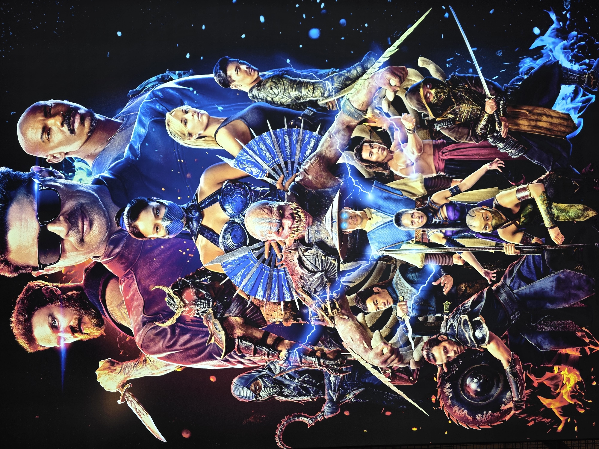 A poster for the movie Mortal Kombat (2021) is displayed, featuring a collage of characters. The background is a black expanse speckled with blue dots. The main characters visible are Jax Briggs, Sonya Blade, Mileena, Shang Tsung, Raiden, Kung Lao, Sub-Zero, Scorpion, Liu Kang, Kano, and an Alien-like creature. Lightning and fire visual effects surround the characters. The location of the poster's placement is not discernible from the image.