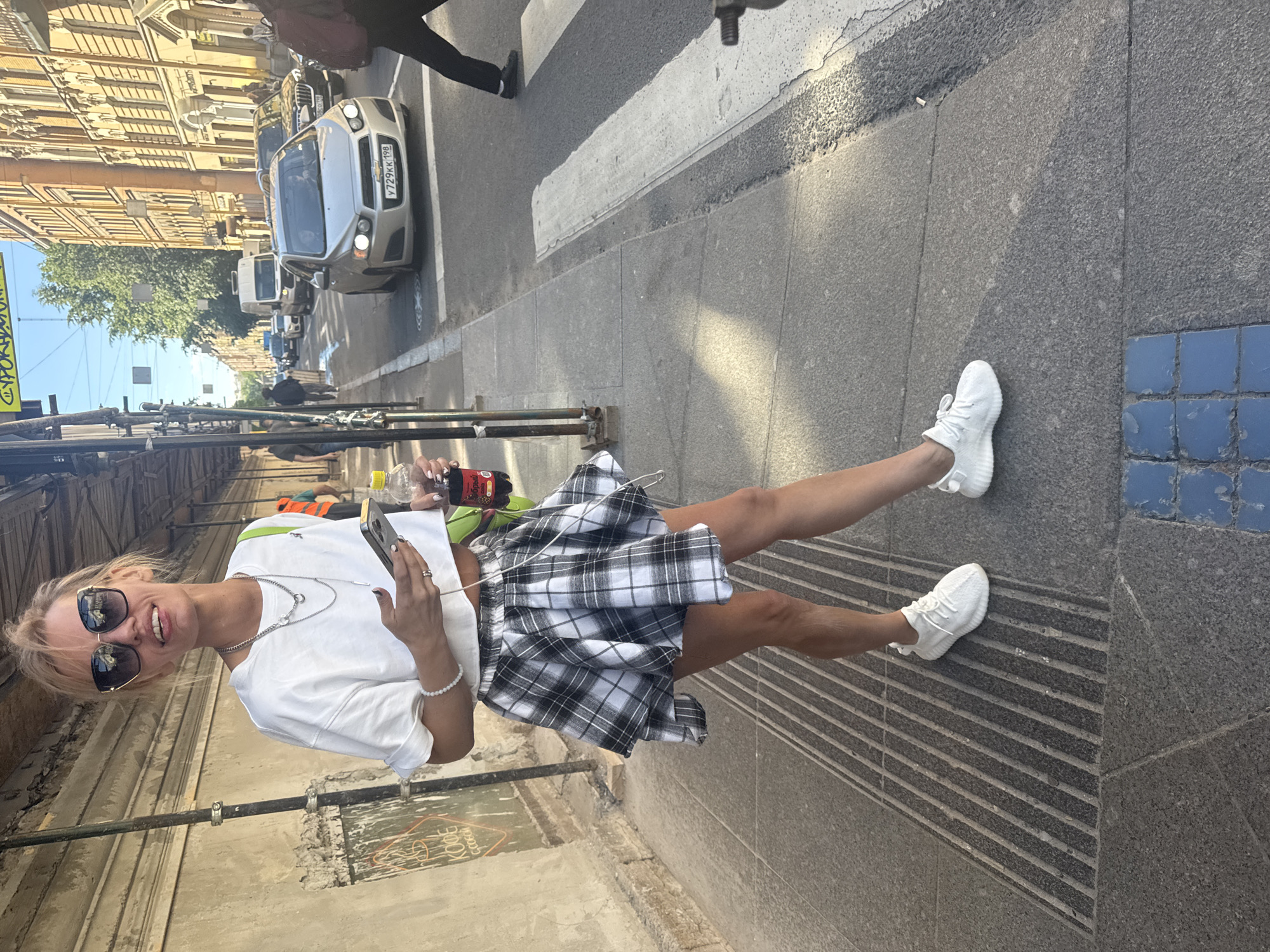 A young blonde woman stands on a city sidewalk in Saint Petersburg, Russia, holding a mobile phone and a bottle of Coca-Cola. She is wearing a white crop top, a black and white plaid skirt, and white sneakers. She has on sunglasses and several necklaces. Behind her, scaffolding lines a building, and across the street, cars are driving on the road, with a pedestrian crossing in the foreground. The sun is shining, casting shadows on the sidewalk.