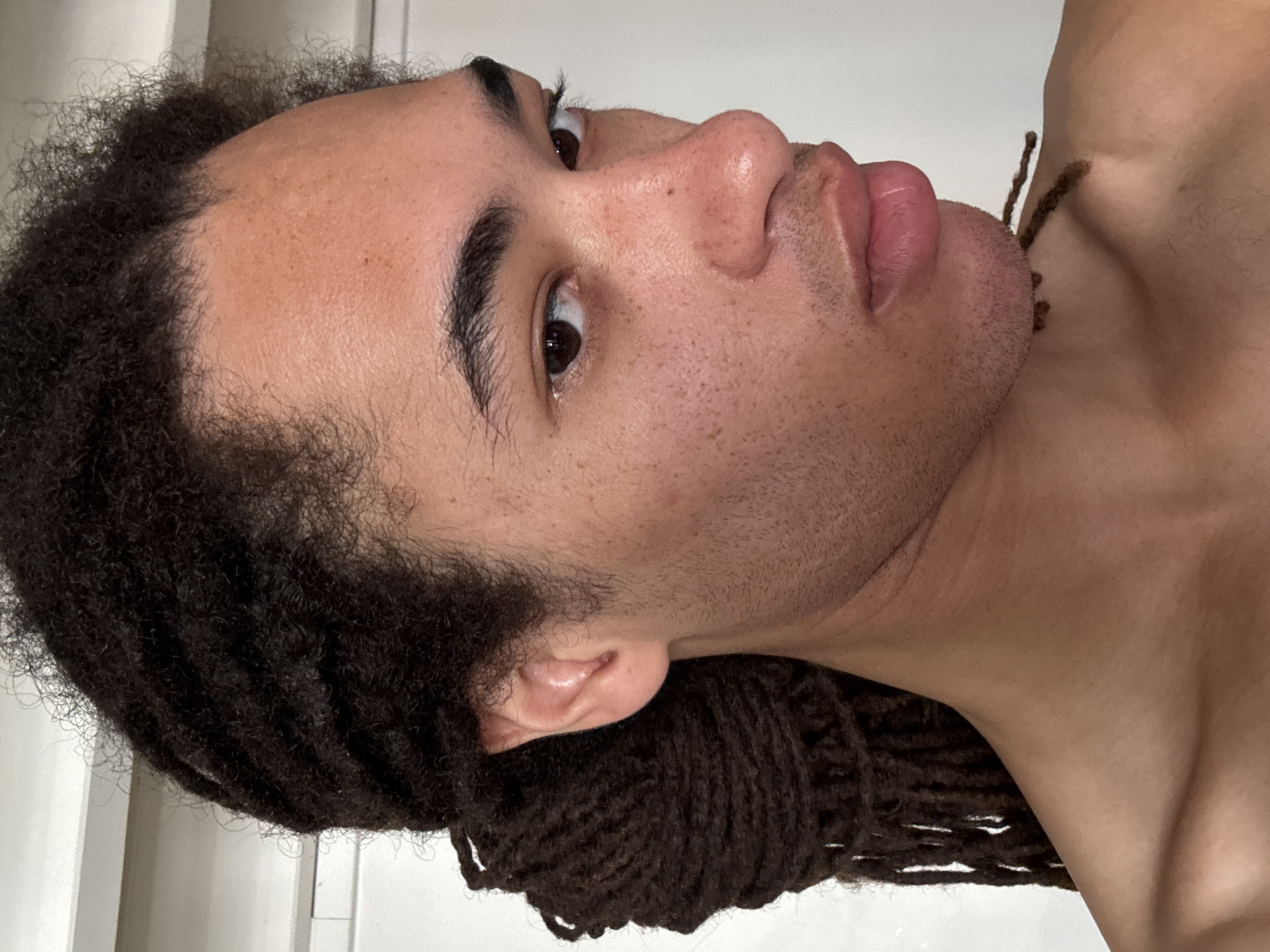 A close-up, slightly angled shot features a person with brown eyes, dark eyebrows, and a tanned complexion. The person has partially dreaded black hair, with some strands resting near their neck. The lips are full and slightly parted. The background is a blurred, light-colored surface, possibly a wall. No indications of the city of Paris, France are visible.