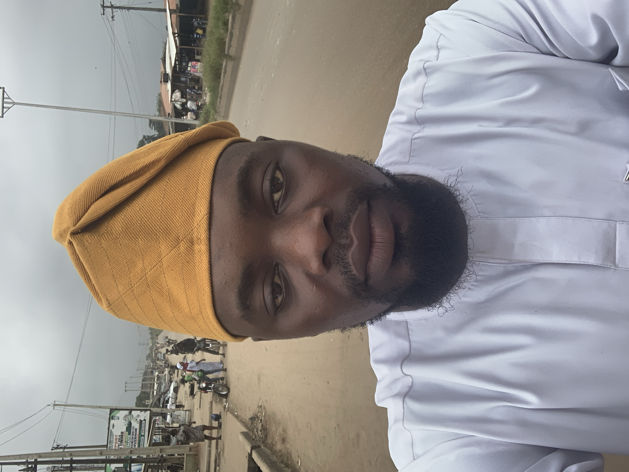 A man, wearing a yellow headwrap and a white garment, takes a selfie. He has dark skin, a beard, and is looking directly at the camera. The background shows a dusty road with several people and motorcycles, suggesting a busy street scene in a town or city. Buildings, some with corrugated iron roofs, line the side of the road. There are also utility poles and power lines visible, indicating an urban or semi-urban environment. A billboard with images and text is also present. The sky appears overcast, suggesting a cloudy day. The image is taken vertically, with the man's head and upper body filling a significant portion of the frame. The overall impression is of a candid snapshot taken outdoors in Shagamu, Nigeria.