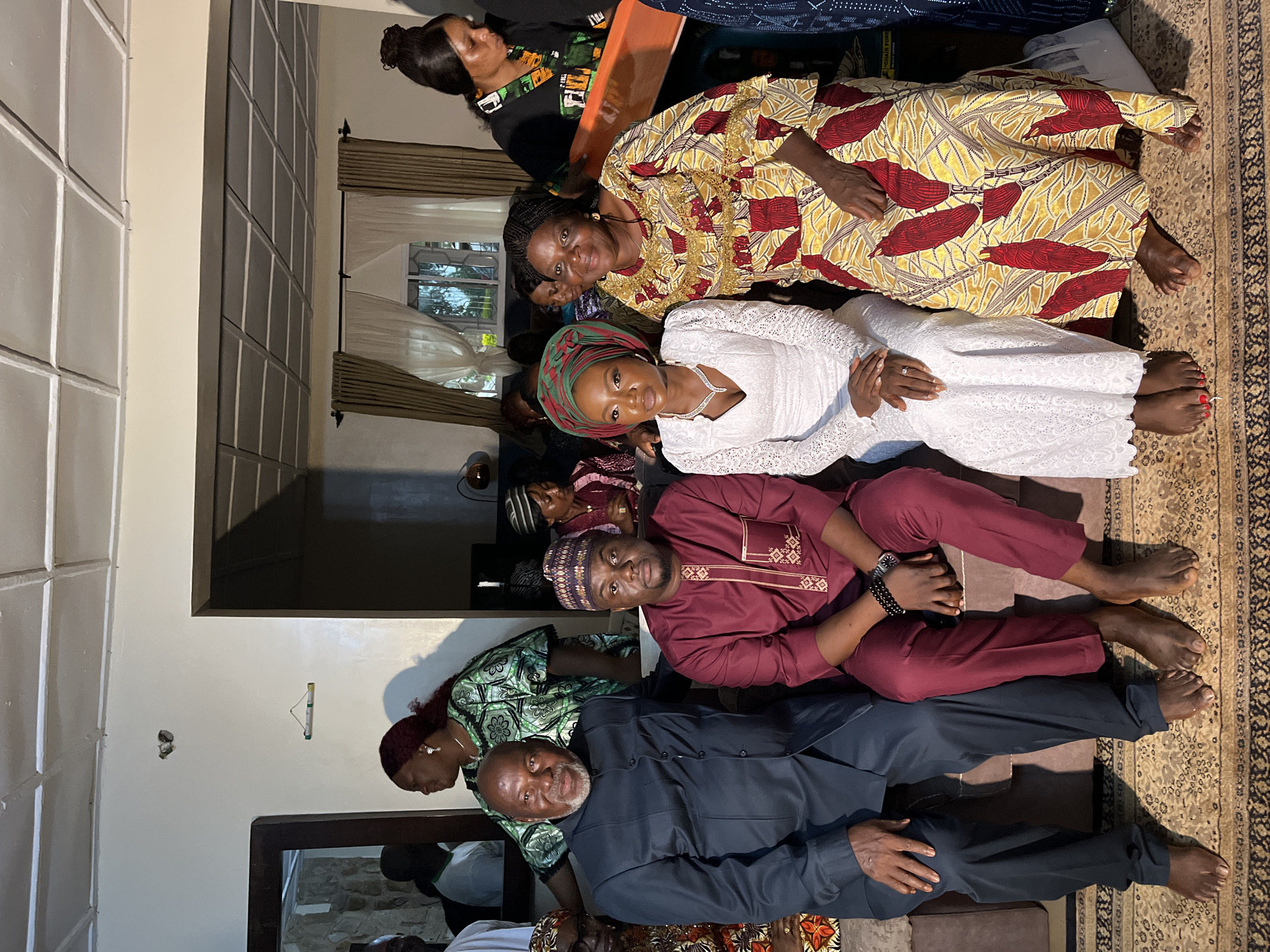 The image depicts an indoor gathering in what appears to be a residential setting, possibly in Bukuru, Nigeria. Five individuals are the primary subjects, seated together.
On the left, an older man with a grey beard and mustache wears a dark blue suit. Next to him, a younger man in a maroon traditional outfit with intricate gold embroidery, including a matching cap, is seated. To the right of the younger man is a woman in a white lace dress, her hair styled up, and wearing a vibrant green and red headscarf. She is adorned with a necklace and earrings. Beside her, another woman is dressed in a yellow and red patterned traditional outfit. A fifth individual, partially visible on the far right, wears a black and green patterned garment.
The group is seated on a brown sofa placed on a patterned rug. The background reveals elements of a home interior, including a doorway, curtains, and what looks like a framed picture on the wall. The lighting suggests an ambient interior light source, with some shadows present. The overall atmosphere seems to be one of a social event or family gathering.