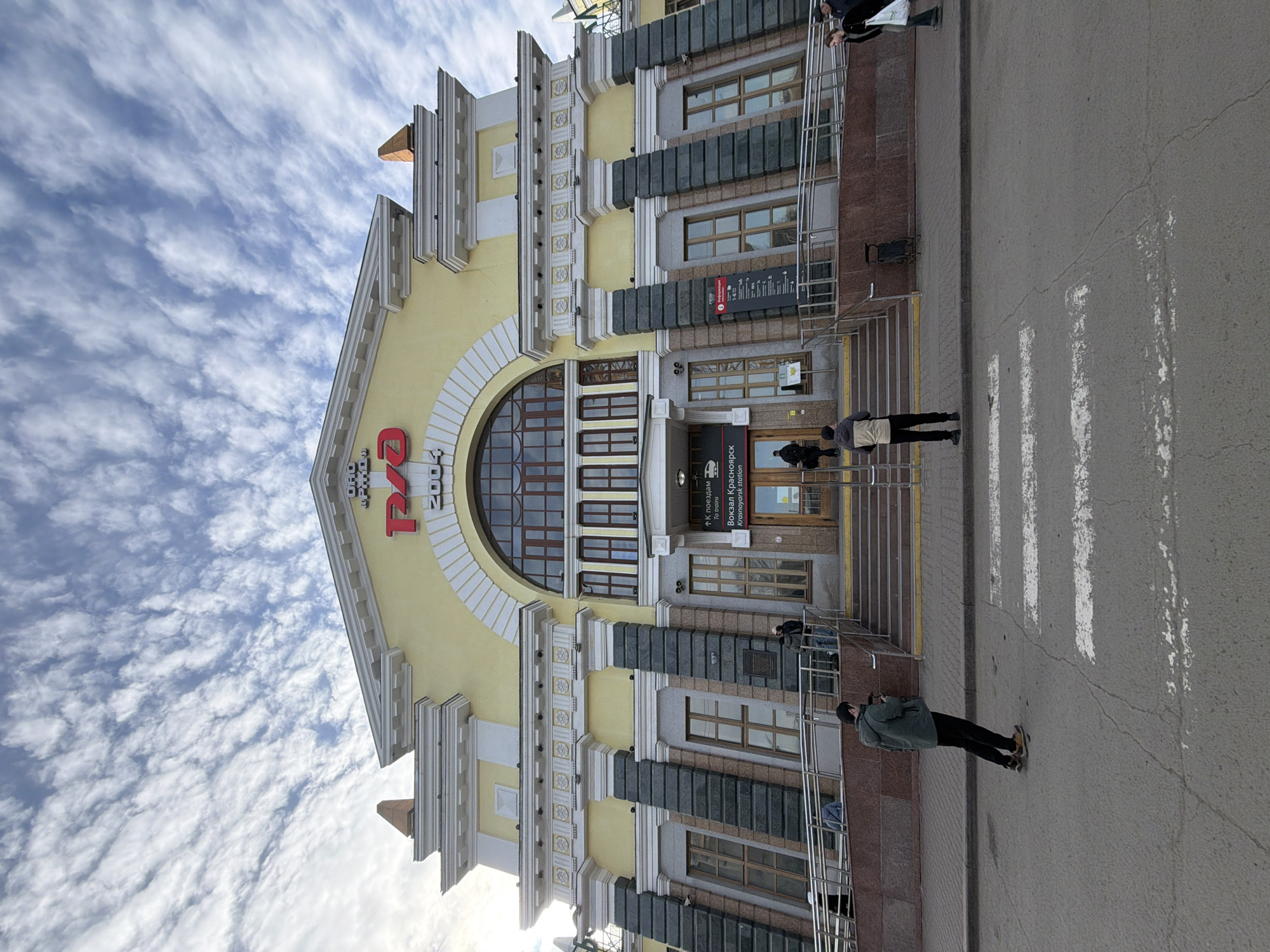 The image shows the Krasnoyarsk railway station in Russia. The building is yellow with white trim and has a large arched window. There are dark grey pillars or pilasters and a sign in Cyrillic above the entrance. There are a few people walking on the sidewalk in front of the building and on the adjacent crosswalk. The sky is blue with scattered clouds.