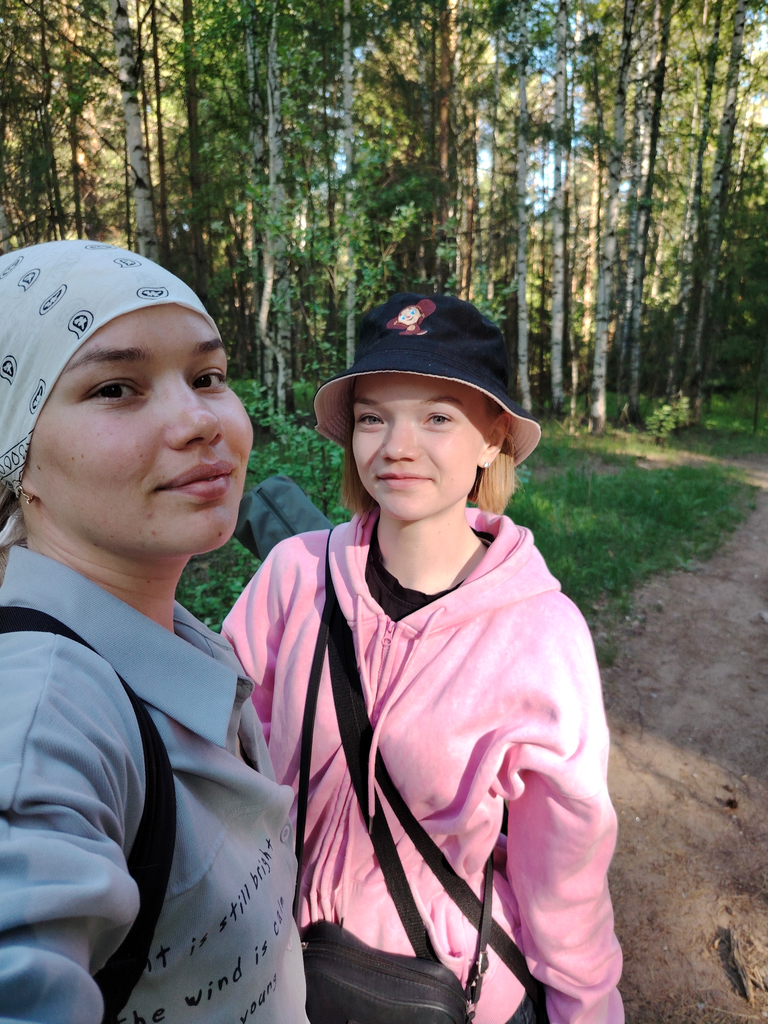 Two young women stand on a dirt path in a birch forest. The woman on the left wears a patterned white bandana, a light grey shirt with text, and a black strap across her chest. The woman on the right wears a black bucket hat with a cartoon figure, a pink hoodie, and a black strap across her chest. The location is near Марковский, Russia.