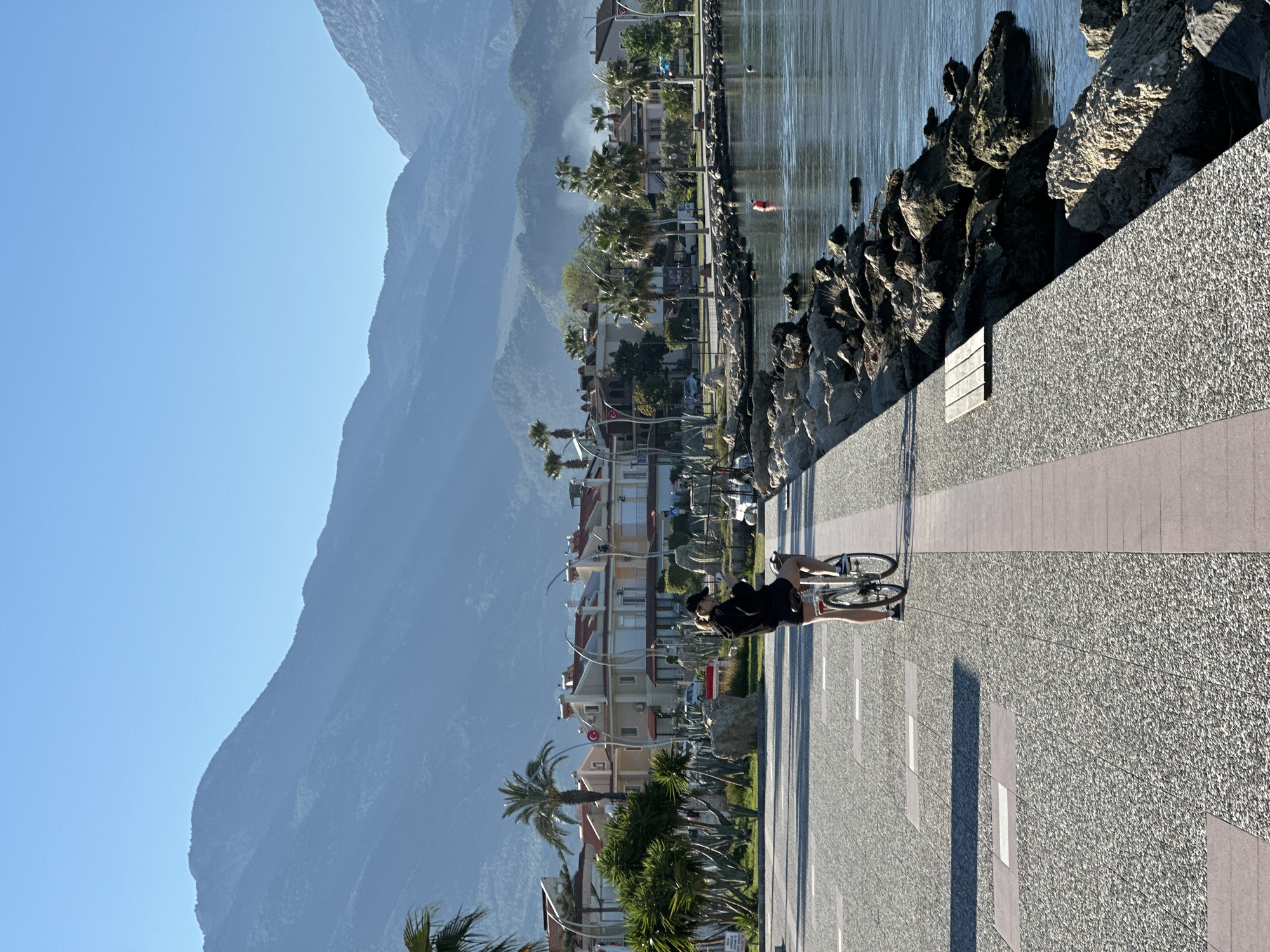The image captures a serene coastal scene in Fethiye, Turkey, under a clear, bright sky, likely in the early morning or late afternoon given the long shadows. A wide, textured promenade with light grey granular paving and darker striped sections runs along the right side of the frame.
In the foreground, a person is seen cycling on the promenade, moving towards the left. The cyclist is dressed in dark attire, including a cap, and is riding a bicycle with visible wheels and frame. Their shadow stretches out long across the pathway.
To the left of the promenade, a barrier of dark, rugged rocks lines the edge of a calm body of water, which reflects the sunlight with gentle ripples. In the mid-distance of the water, a small red object is barely visible, possibly a buoy or a distant swimmer.
Beyond the promenade, a row of buildings with light-colored facades and reddish-orange tiled roofs extends along the coast, interspersed with numerous lush palm trees and other green foliage. Streetlights are also visible along this urban stretch.
The background is dominated by large, impressive mountains that rise sharply, their slopes appearing somewhat hazy, indicating their grandeur and distance. The sky above is a vibrant, clear blue. The overall atmosphere is peaceful and scenic, showcasing a pleasant environment ideal for leisure activities like cycling. No discernible text is visible within the image.