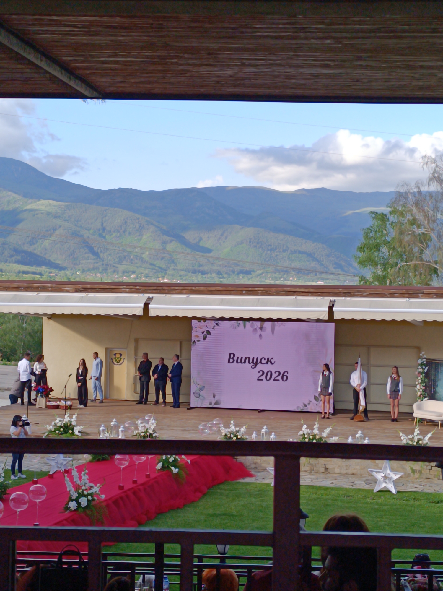 The image shows an outdoor graduation ceremony in Chelopech, Bulgaria, likely in 2026, based on the stage backdrop. A group of people stands on a stage decorated with white flowers and a red carpet. The backdrop displays "Випуск 2026". Students in school uniforms are present on the stage. Mountains and a cloudy sky are visible in the background. A camera operator is documenting the event. The scene is viewed from behind a railing, with audience members visible in the foreground.