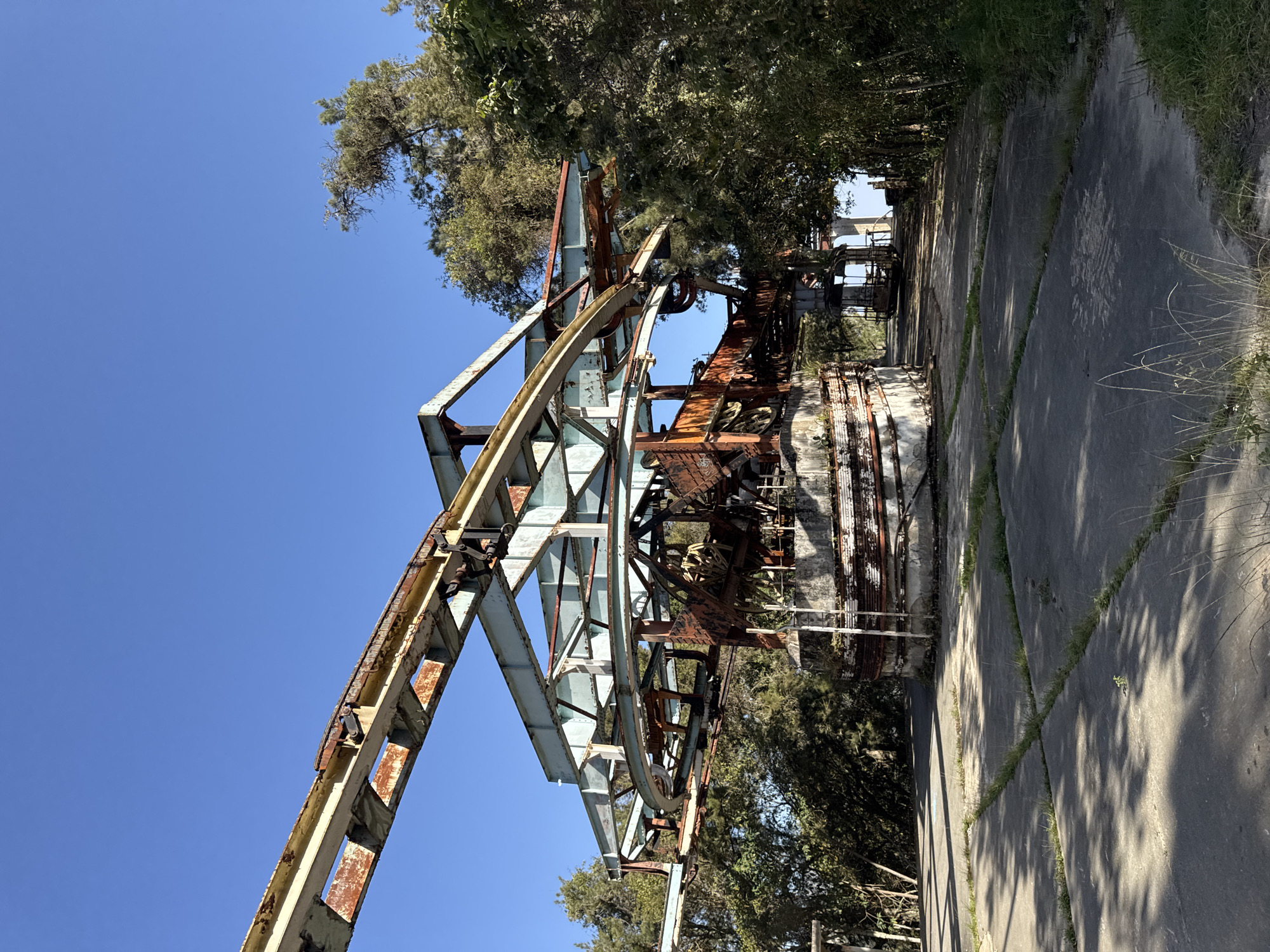 This image depicts the remnants of an abandoned, rusted metal structure in San Antonio de Galipán, Venezuela. The structure is complex, appearing to be an industrial-type construction with a skeletal framework of girders, supports, and circular elements. Rust is pervasive, indicating significant weathering and disuse. The structure is surrounded by trees and foliage, and it stands on a concrete foundation or surface with cracks and vegetation growing in the seams. The scene is bathed in sunlight, with a clear blue sky in the background. There are no people visible in the image. The focus is on the decay and abandonment of this once-functional structure.