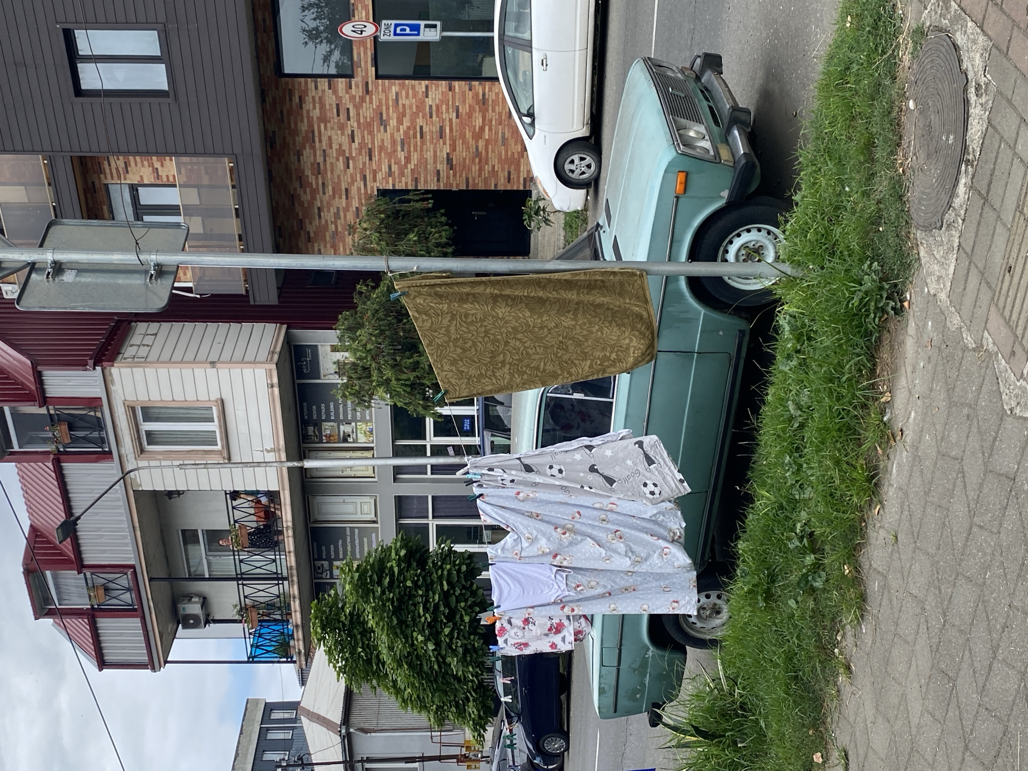 A teal colored vintage car is parked on a city street in Batumi, Georgia. The car is partially obscured by laundry hanging on a clothesline stretched between a pole and the car. The laundry includes a white t-shirt and patterned sheets. A large, olive-green tapestry hangs from the same clothesline. The street is lined with buildings, one of which is under construction with exposed brickwork. A speed limit sign with the number 40 is visible on a pole in the background. The ground is paved with cobblestones and there is a patch of overgrown grass on the side of the road. The sky is overcast.