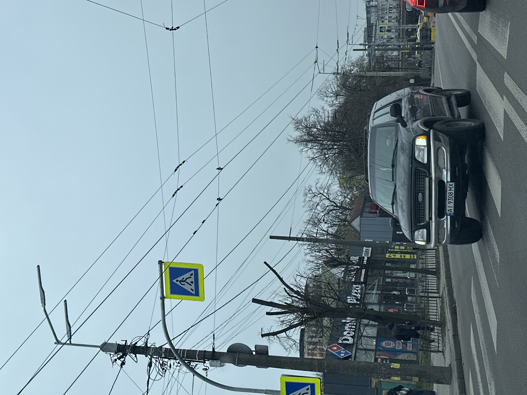 The image shows a street scene in Kyiv, Ukraine, with a black Ford SUV in the foreground. The vehicle's license plate reads "AI 9308 MX". The SUV is on a zebra crossing. To the left of the SUV and across the street is a Domino's Pizza store, visible through bare tree branches and a metal fence. Pedestrian crossing signs are present, one is blue and yellow. Above, numerous power lines crisscross the sky. Two people are seated on a bench near the Domino's Pizza. The weather is overcast, creating a muted light across the scene. Other buildings are visible in the background.