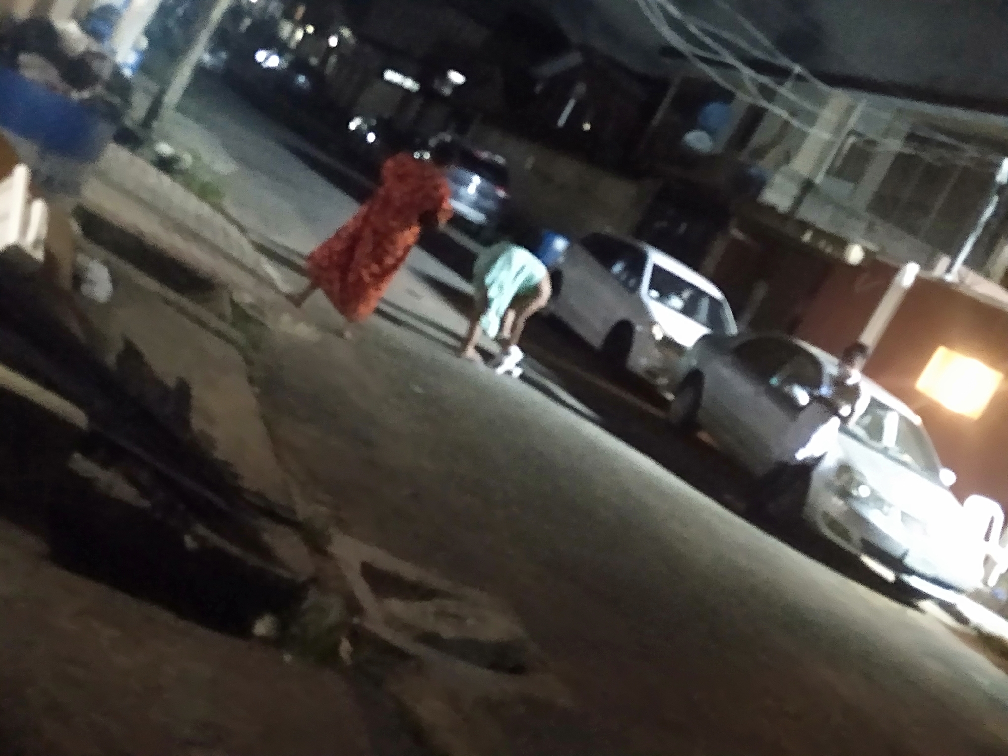 The image shows an urban scene at night. Several parked cars line a street with buildings visible in the background. In the foreground, a person in a red dress is standing, while another person wearing a light-colored top is bending over. The location is the city of Unknown, Unknown.