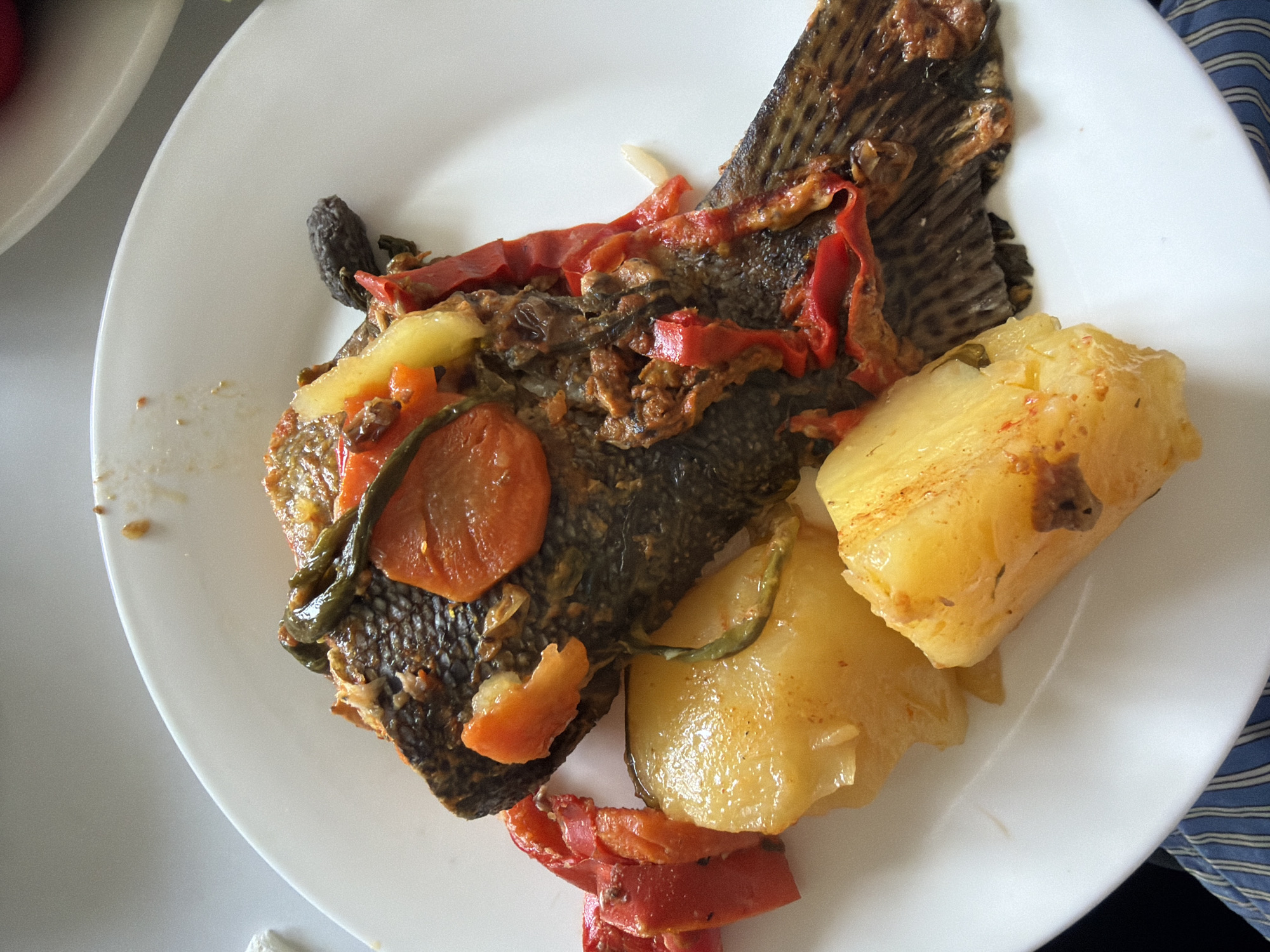 On a white plate sits a cooked fish, skin-on and garnished with red peppers, carrots, and green beans. Next to the fish are several yellow potatoes. The plate rests on a white surface, with a blue and white striped fabric visible on the right. The image was likely taken in Yerevan, Armenia.