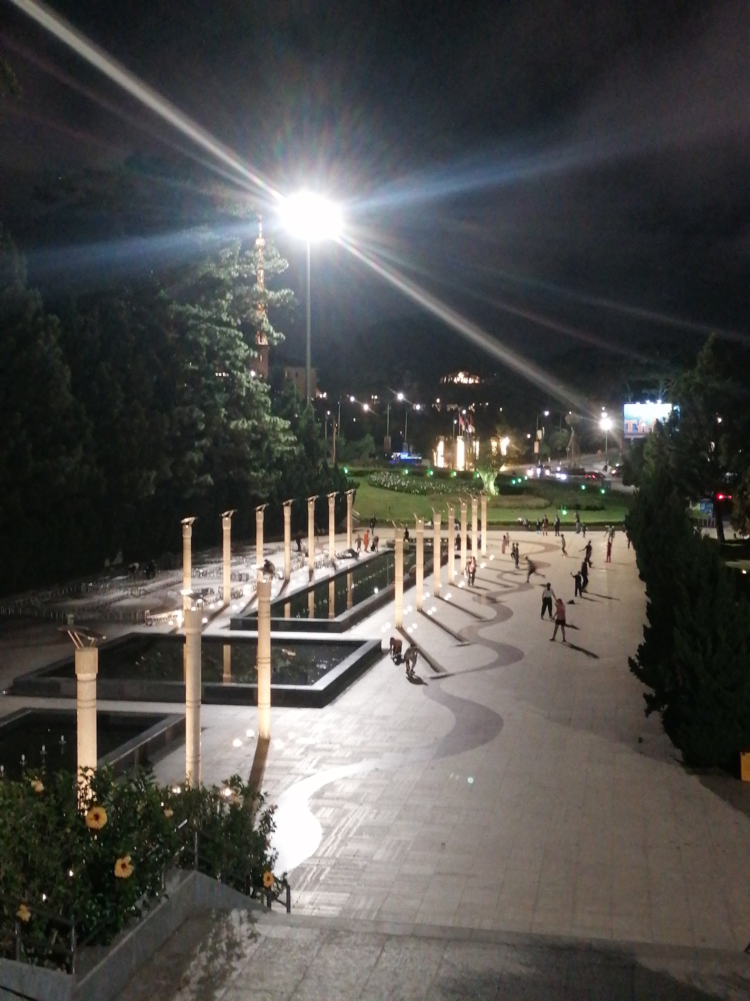 The image shows a nighttime scene in Da Lat, Vietnam. The foreground features a multi-tiered fountain area with rectangular pools lined by tall, cylindrical pillars capped with horizontal, light-reflecting surfaces. A winding, paved walkway extends from the fountain area towards a street lined with lights in the background. Many people are walking, standing, and milling about on the walkway. The scene is illuminated by a very bright street lamp with visible lens flare, other streetlights, and the glow of buildings in the background. To the left and right of the frame are dark trees and foliage. In the background, buildings are visible atop a hill. A large billboard is located at the end of the walkway. Yellow flowers are visible in the lower left foreground near a short flight of stairs.