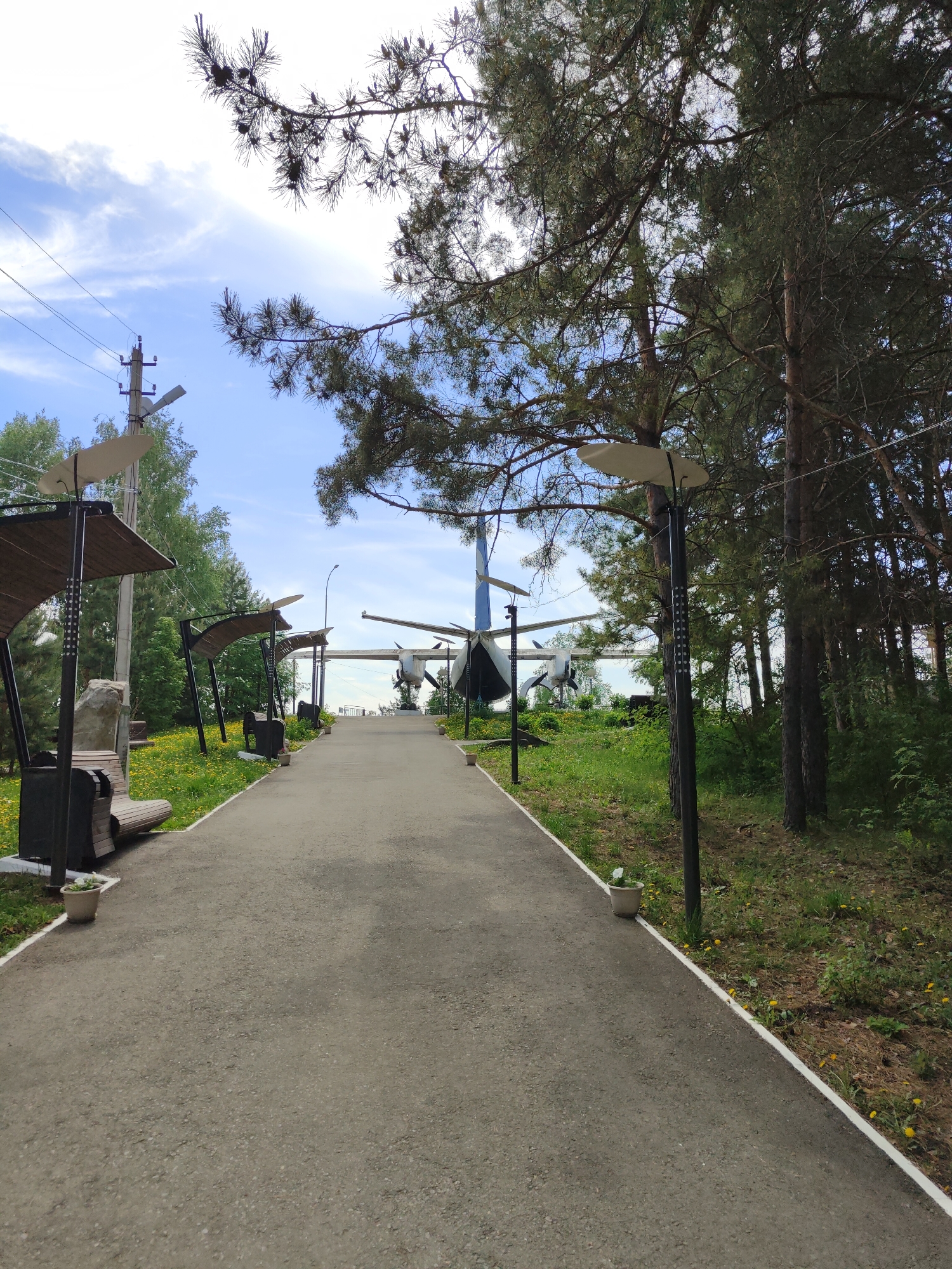 The image depicts a paved walkway leading towards a plane in a park or outdoor museum. Benches and shaded seating areas line the left side of the walkway, while a line of trees and lamp posts are on the right. The sky is visible in the background. The location is an outdoor area, potentially a park or museum, in the city of Unknown, Unknown.