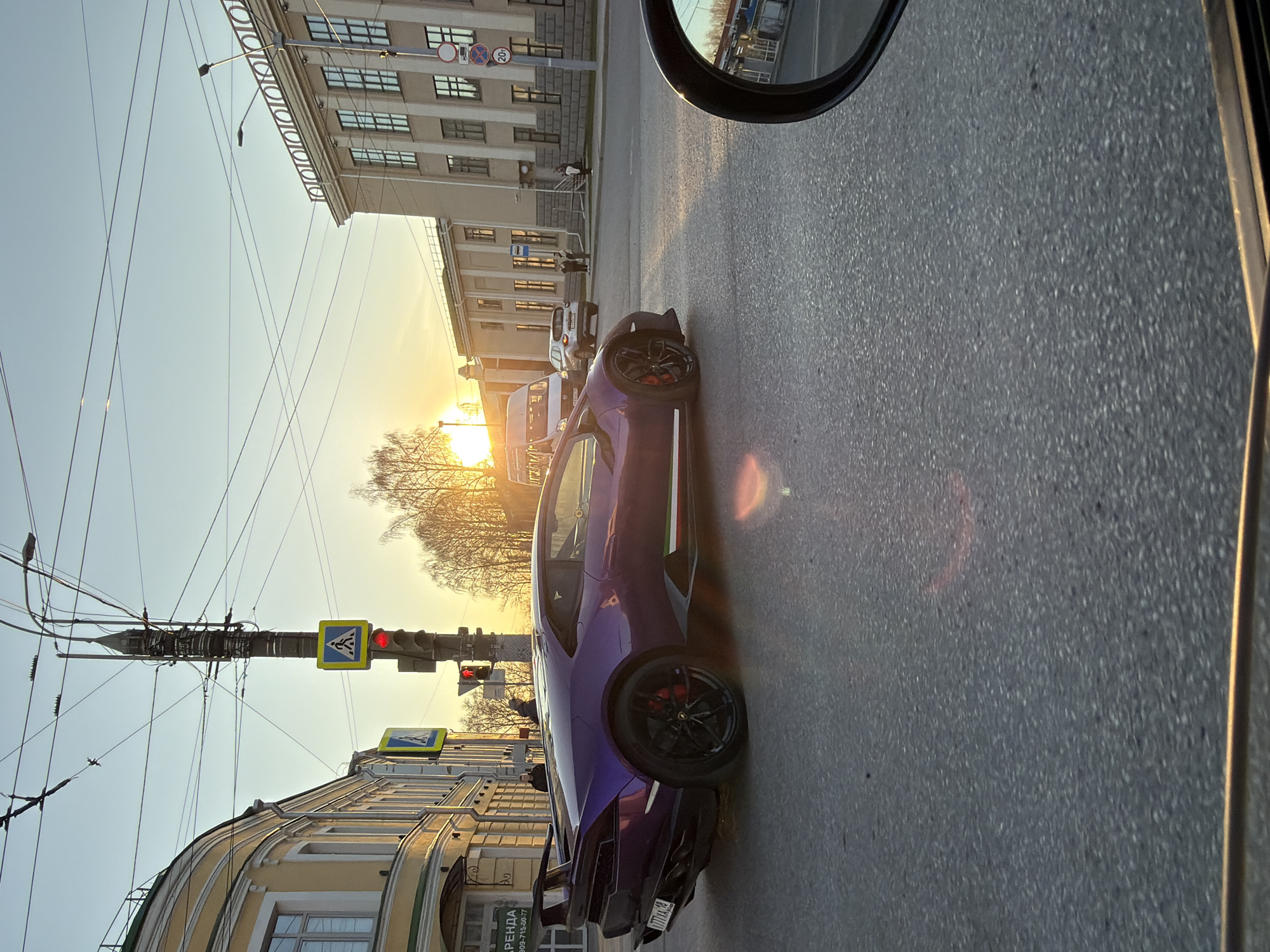 The image captures a street scene at sunset in Izhevsk, Russia. The main subject is a purple Lamborghini parked on the right side of the road. The car's rear license plate reads "777XA77". A traffic light shows red, and pedestrian crossing signs are visible on a utility pole. Buildings line both sides of the street: a beige two-story building with arches on the left, and a larger, lighter-colored building with multiple windows in the background. The sun is setting behind trees. A car's side mirror is visible in the upper right corner. Several people are standing near the building in the background. The street is paved with asphalt.