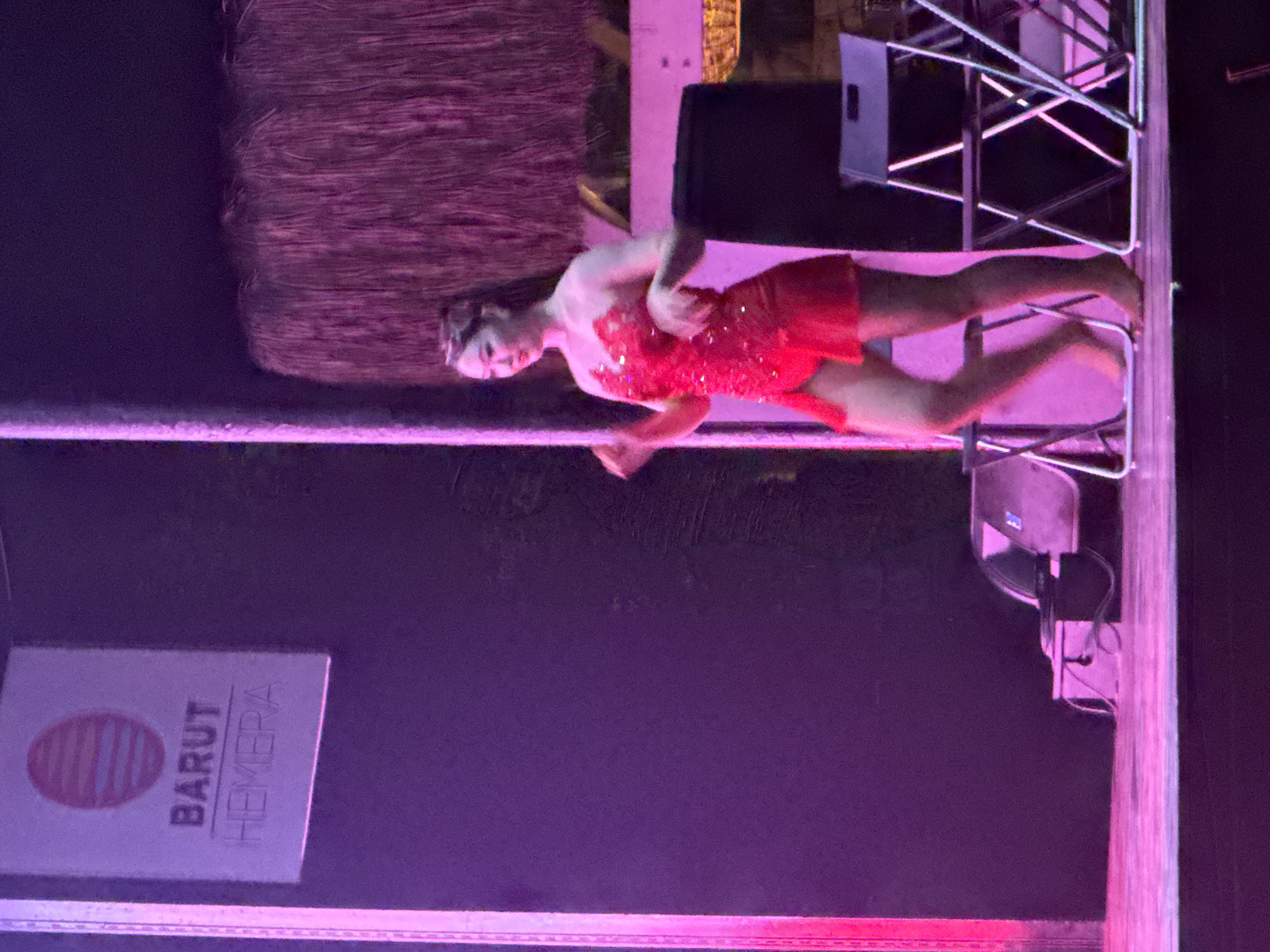 A woman in a short, red, sequined dress is dancing on a stage. The stage is lit with pink and purple lights. Behind the woman, there is a wall with a sign that reads "BARUT HEMERA". There are also some chairs and speakers on the stage. The scene appears to be from a performance at the Barut Hemera hotel in Manavgat, Turkey.