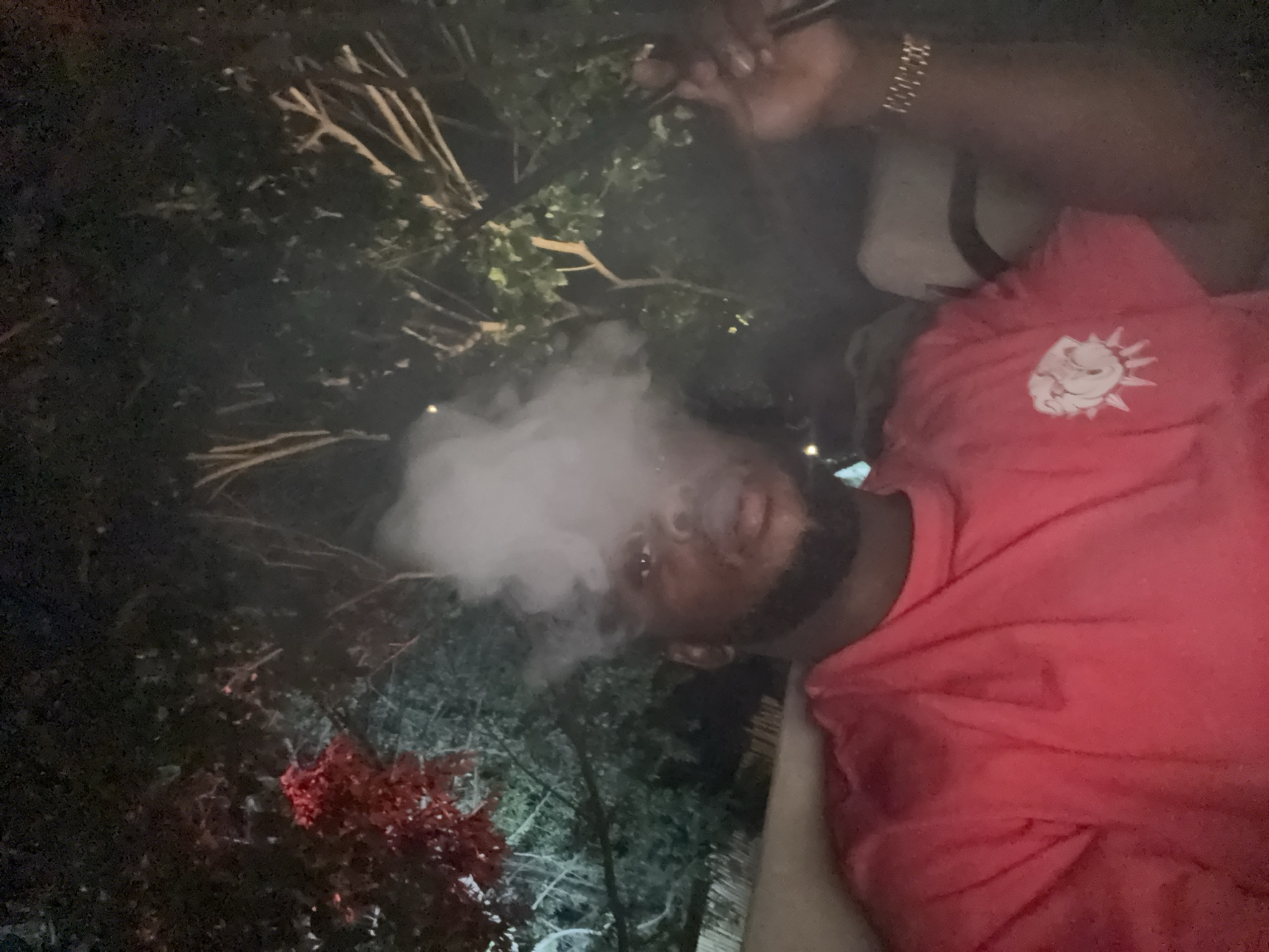 A man with dark skin and a beard, wearing a coral-colored t-shirt with a graphic print, exhales a cloud of white smoke. He holds an object in his right hand. He wears a silver bracelet. The backdrop consists of dark foliage, trees, and what appears to be a nighttime sky. Location details for Abuja, Nigeria are not discernible from the visual information.