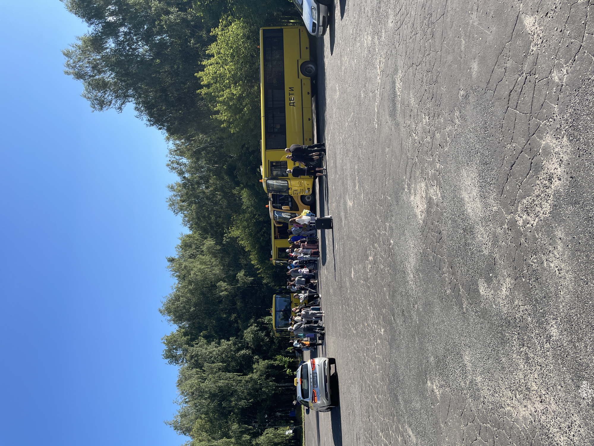 In Salihorsk, Belarus, on a bright sunny day, a group of people, including children, are gathered in a parking lot near several yellow buses and a silver taxi. The buses, one of which has the word "ДЕТИ" (Children) written on its side, appear to be parked in a line. Some individuals are standing near the buses, while others are with luggage. The taxi is parked on the left side of the frame. The environment includes a paved surface with visible cracks and scattered gravel, and a backdrop of lush green trees under a clear blue sky.