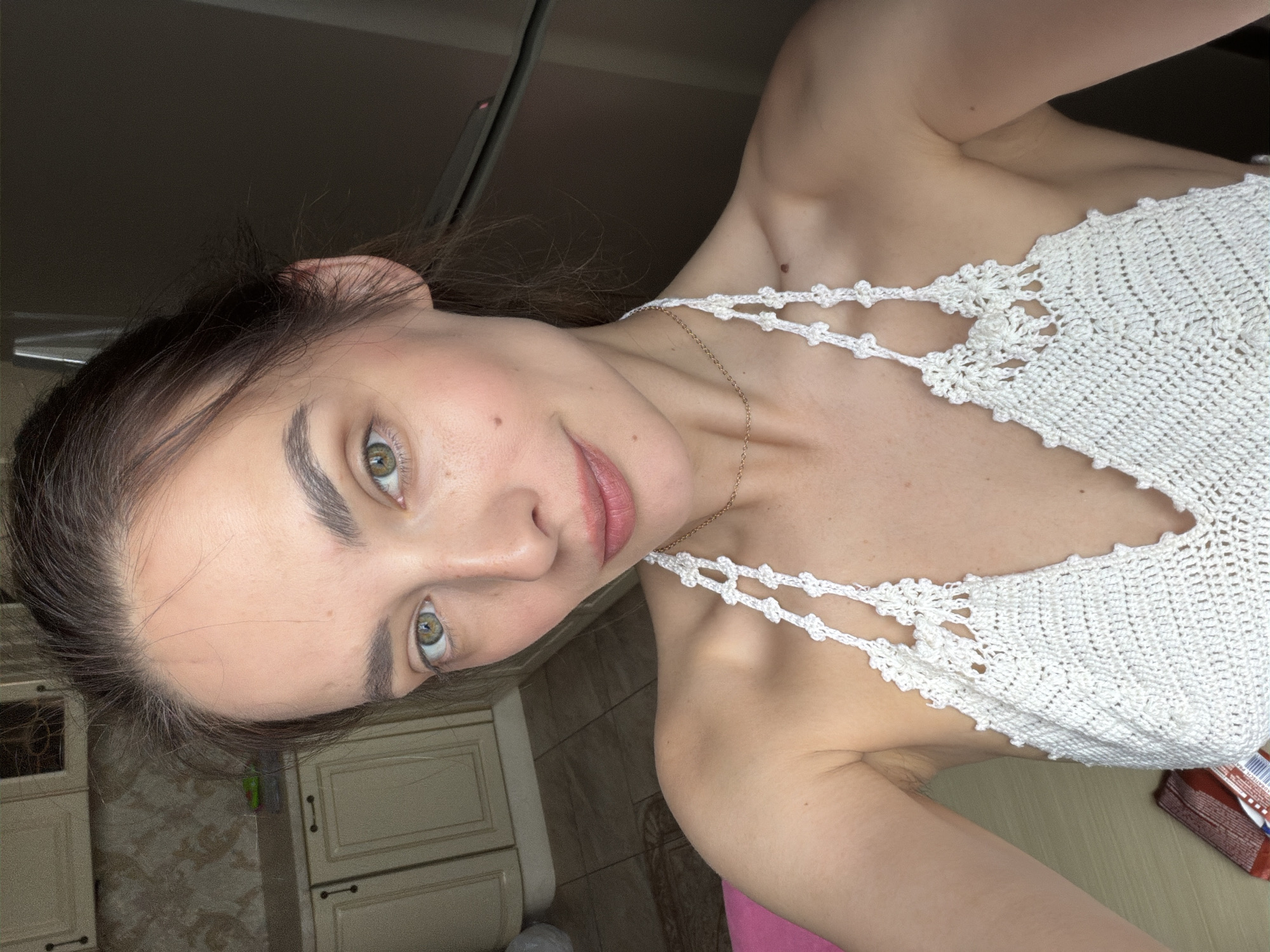 A young woman with green eyes and dark brown hair is taking a selfie in what appears to be a kitchen. She is wearing a white crochet halter top. The background shows kitchen cabinets and a tiled floor. There are no other people or obvious signs of urban environment in the visible area. The exact location within Ufa, Russia cannot be determined from the image.
