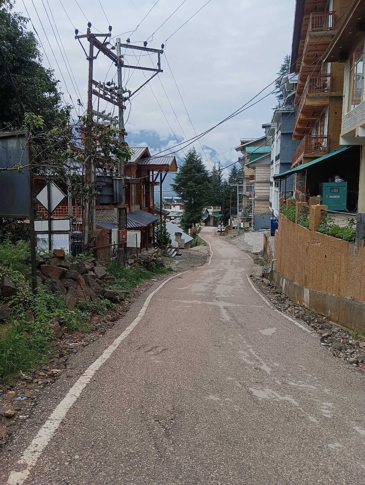 This image depicts a street scene in the city of Chhyal, India. The road is paved with asphalt and has a white painted line on the left side. On the left, there are piles of rocks and green vegetation. A utility pole with numerous wires and a transformer is also visible. Several buildings with wooden balconies and balconies with railings line the right side of the street. In the background, mountains are visible, obscured by clouds and trees. The sky is overcast. A green generator is visible on the far right of the image.