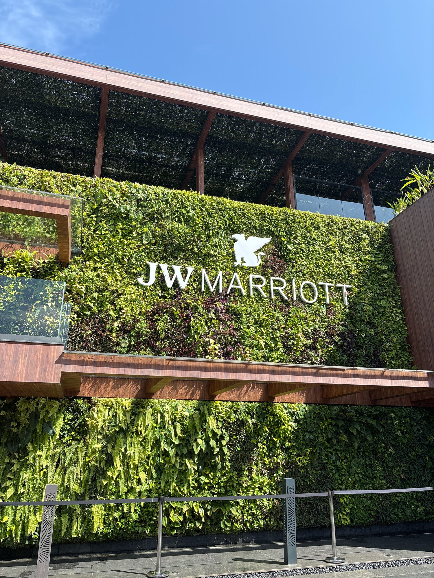 The image shows the exterior of the JW Marriott hotel in Vagator, India. A large, living green wall covers the facade of the building, with the hotel's name and logo prominently displayed in white lettering. The logo features a white bird silhouette above the text "JW MARRIOTT". The green wall is made up of various plants, including ferns and other leafy foliage. A wooden overhang supported by wooden beams is visible above the green wall. Below the green wall, a pathway is lined with dark grey paving stones and a decorative border of small black stones. Two stanchions with a grey retractable belt are placed on the pathway, suggesting a queueing or crowd control system. The sky above is clear and blue.