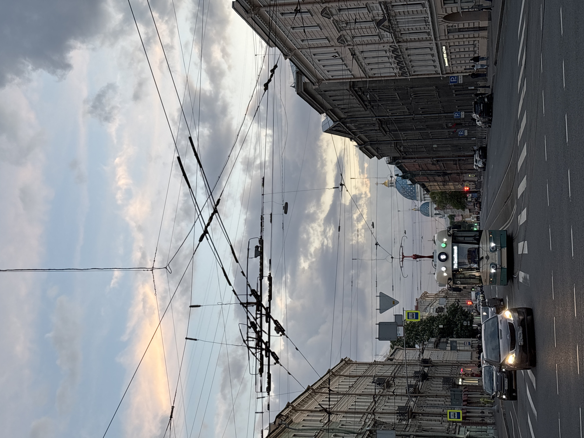 This eye-level, vertical street view captures an urban scene in Saint Petersburg, Russia. A complex network of trolleybus overhead wires crisscrosses prominently across the upper and central portions of the frame against a cloudy sky. The sky displays varying shades of grey and pale blue, with hints of soft orange and yellow light peeking through, suggesting either dawn or dusk.
The street, paved with asphalt, occupies the right side of the image, featuring white lane markings and a pedestrian crossing. In the mid-ground, a modern green and white trolleybus with illuminated headlights approaches. Its route number "10" is visible on the front display. A dark-colored car, possibly a sedan or SUV, is closer in the foreground, also with its headlights on. Its Russian license plate, "124 HAX 47", is clearly visible, with "47" indicating the Leningrad Oblast region. Several other cars are parked or moving further down the street.
Multi-story historical buildings with classical architectural details line both sides of the street. These ornate, light-colored structures feature numerous windows and intricate cornices, typical of Saint Petersburg. A few indistinct figures are visible on the sidewalks, near parked cars. Multiple blue 'P' signs, indicating parking zones, are visible along the street.
In the background, beyond the buildings, the distinctive domes of Saint Petersburg's cathedrals are visible. A large blue dome adorned with white stars, characteristic of the Kazan Cathedral, is prominent, along with at least one other golden dome, underscoring the city's unique architectural heritage. The overall atmosphere is that of a calm urban street during the transitions of day.