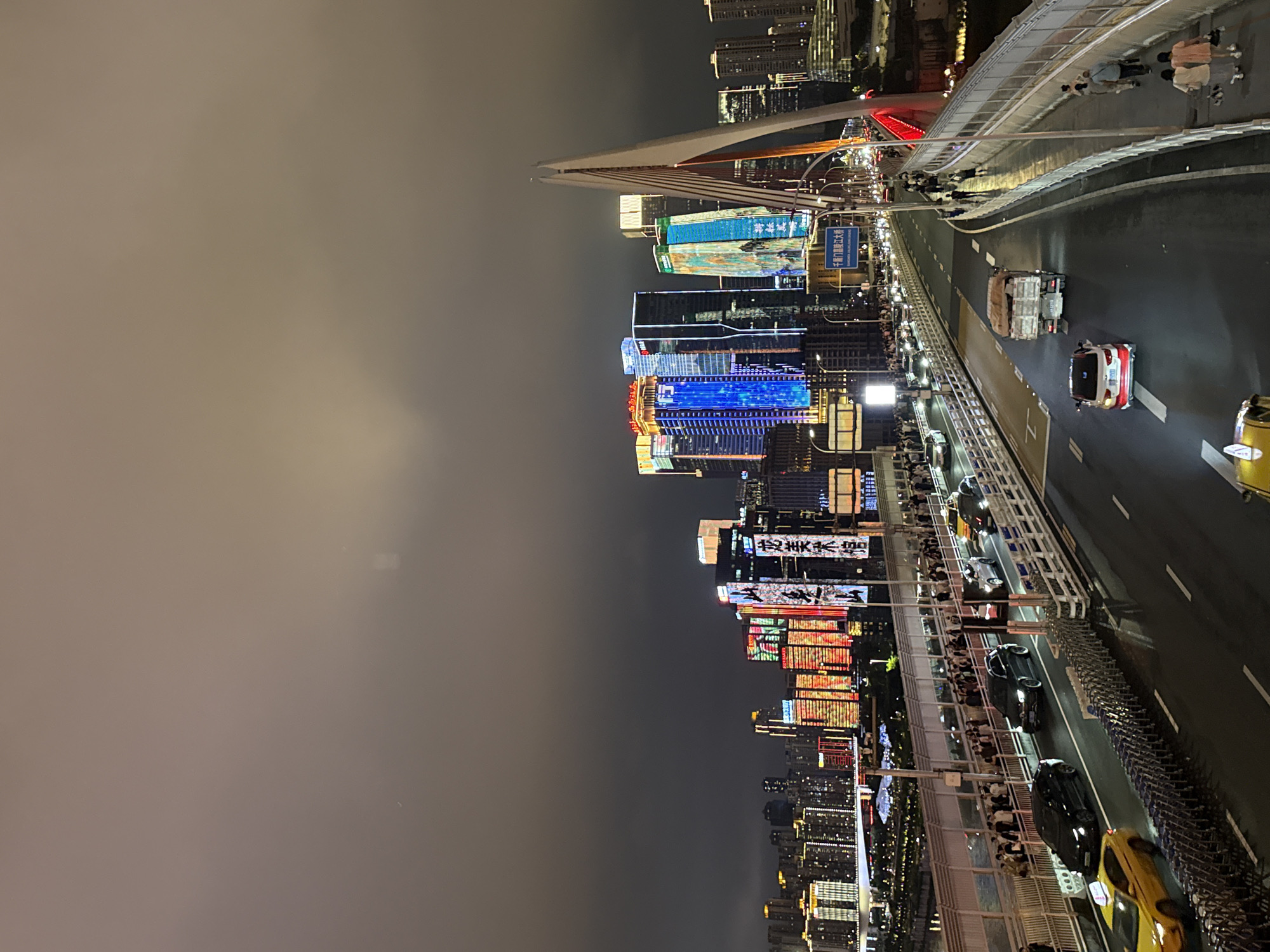 This is a night-time, high-angle, wide shot of a bridge in Yuzhong District, China, with a city skyline illuminated by bright lights in the background. The bridge has multiple lanes of traffic moving in both directions, with cars and a truck visible. A pedestrian walkway runs alongside the traffic lanes, with numerous people walking and standing. The city skyline features tall, modern buildings adorned with colorful LED lights displaying various patterns and Chinese characters. A prominent, modern cable-stayed bridge structure is visible on the right side of the frame. The sky is dark and cloudy, obscuring any stars or moon.