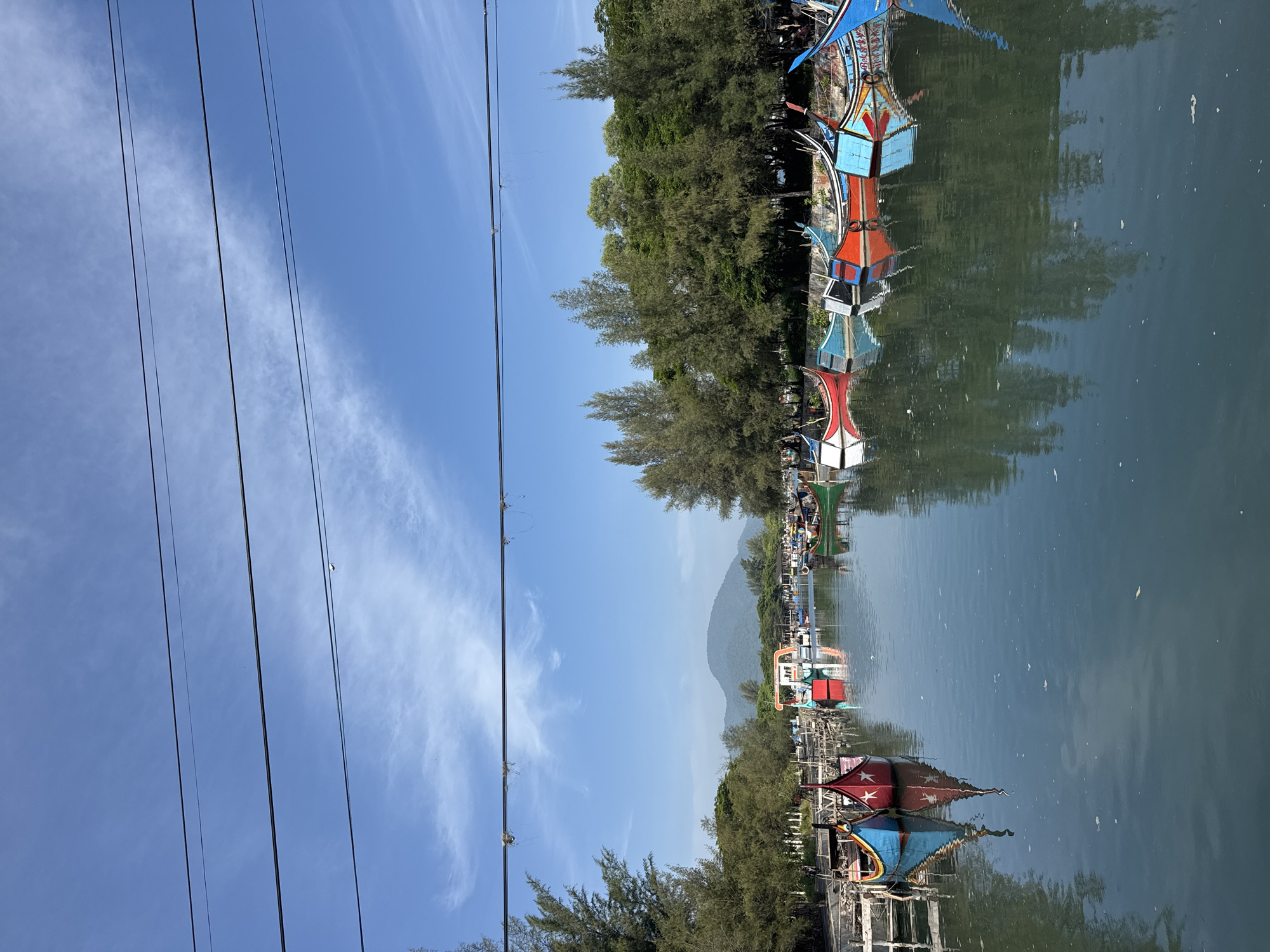 A photograph shows a row of colorful fishing boats docked in a canal in Banda Aceh, Indonesia. The boats are painted in vibrant hues of red, blue, green, and yellow, with intricate designs and flags. They are moored alongside a wooden jetty, with lush green trees and a distant mountain range forming the backdrop. The sky is a clear blue, with a few wispy clouds. The water in the canal is calm and reflects the boats and surrounding scenery. A series of overhead power lines are visible in the foreground, crossing the frame horizontally. The scene evokes a sense of tranquility and offers a glimpse into the local maritime culture of Banda Aceh.