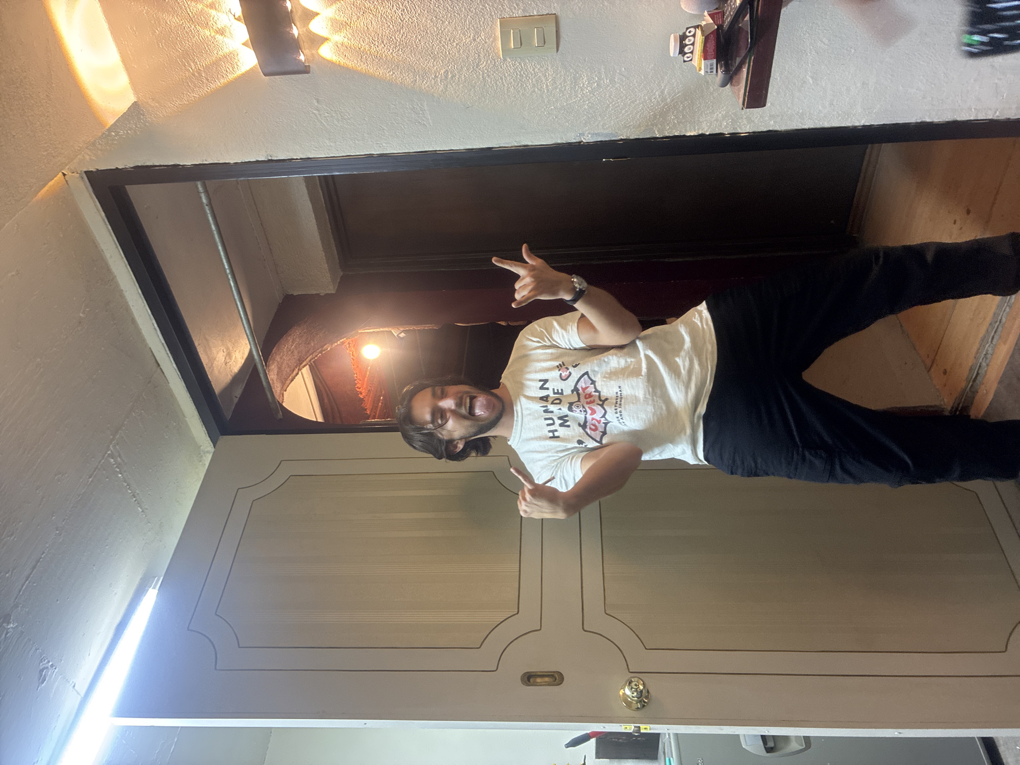 A man is standing in a doorway, facing the camera and throwing a "rock on" hand gesture with both hands. He is wearing a white t-shirt with black lettering, and black pants. The doorway leads into another room with a dark-colored door. The adjacent wall is painted with a light color, with a light fixture near the top of the wall. A light switch is also visible. The location appears to be indoors. I am unable to determine the location is in the city of Huixquilucan de Degollado, Mexico.