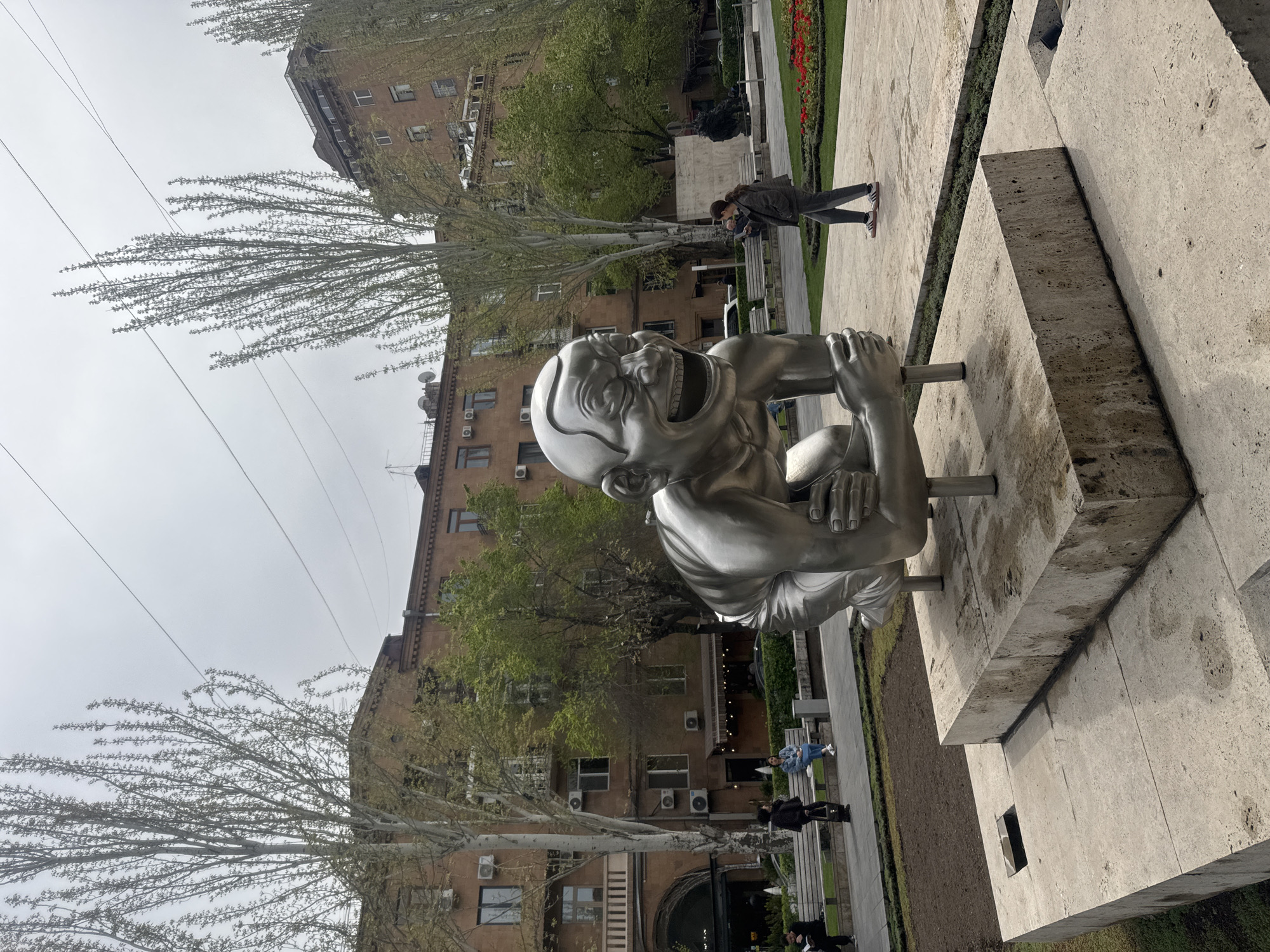 The image depicts the "Men" statues by David Yerevantsi in Yerevan, Armenia. A large, silver-colored statue of a laughing man is the prominent subject. The statue is situated on a concrete base with textured surfaces. People are walking in the background, along a park walkway lined with benches and grass. Buildings stand in the background, indicating an urban setting. The sky is overcast. Trees are scattered throughout, with branches displaying early signs of budding. Power lines are visible against the sky. The statue is located in the Cascade complex.