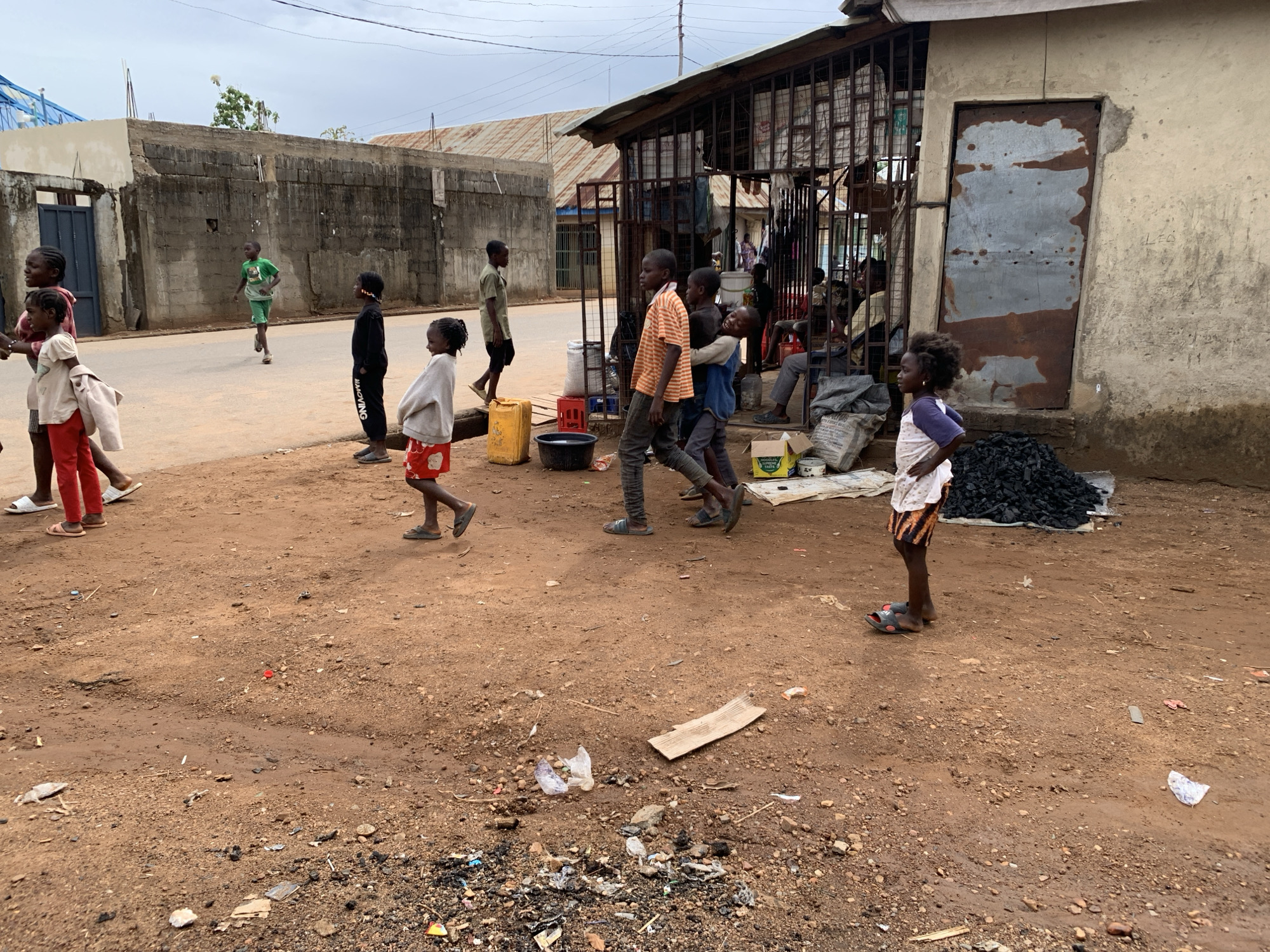 Location: A street scene in Akwanga, Nigeria.
People: A group of children are present, some walking and some standing. Several adults are seated inside a caged shop.
Objects: A concrete wall, a metal-roofed building, plastic containers, debris, a bag of charcoal, and cardboard are visible.
Actions: Children are walking and standing on a dirt road. Adults are sitting. A child is running down the street.
Environment: The environment appears to be a poor, underdeveloped area with a dirt road and simple buildings.