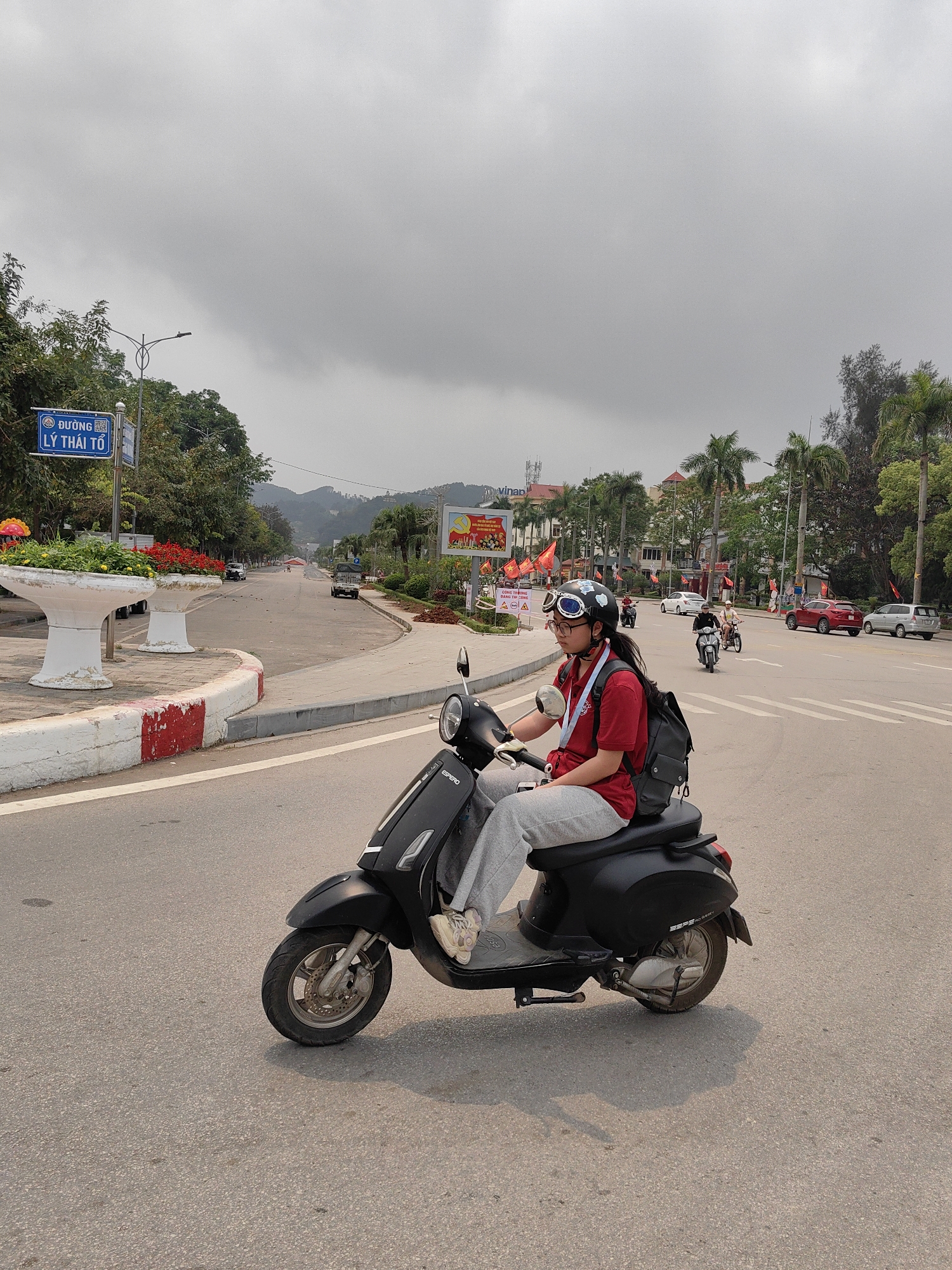 A medium shot captures a young person riding a black Espera scooter on a city street in Lương Văn Tri, Vietnam. The individual, wearing a red shirt, gray sweatpants, white sneakers, a black helmet, and aviator goggles, also has a black backpack. The street features a curved median with planters, red and white painted curbs, and a blue street sign reading "Đường Lý Thái Tổ" (Ly Thai To Street). Palm trees line the street further in the distance, and several other scooters and vehicles are present. A banner advertising "Vinap" is visible in the background, and overcast skies fill the upper portion of the frame.