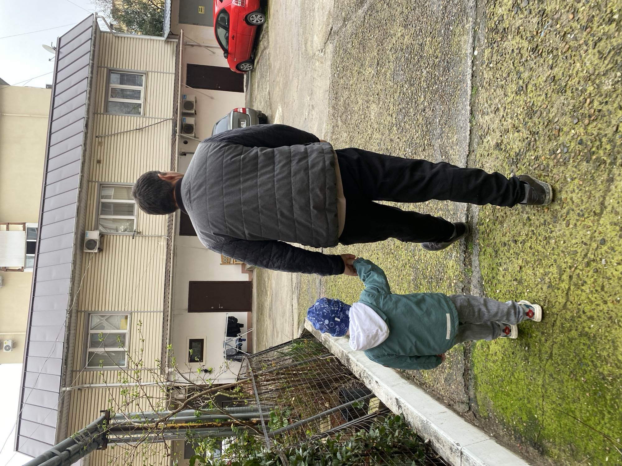 A man and a child are walking hand-in-hand on a paved surface next to a low wall and vegetation. The man is wearing black pants, dark shoes, and a gray vest. The child is wearing gray pants, a green jacket with a white hood, and a patterned blue hat. They are located in an urban environment, with buildings visible in the background, including one with a red car parked in front of it. Based on the architectural style and vegetation visible, the scene is plausibly located in Sochi, Russia.