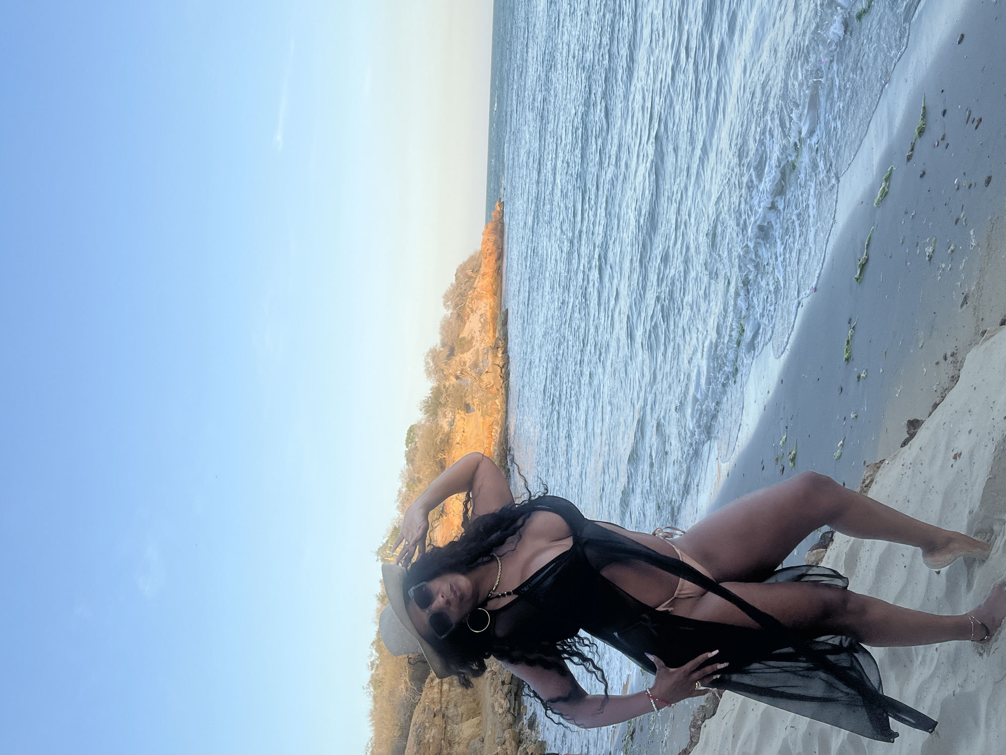 A woman lies on the beach in El Yaque, Venezuela. The sky is clear and blue. The ocean waves lap against the shore. The woman wears a black cover-up, a swimsuit, sunglasses, and a hat. She is posing for a photograph. In the background, there are rock formations and vegetation.