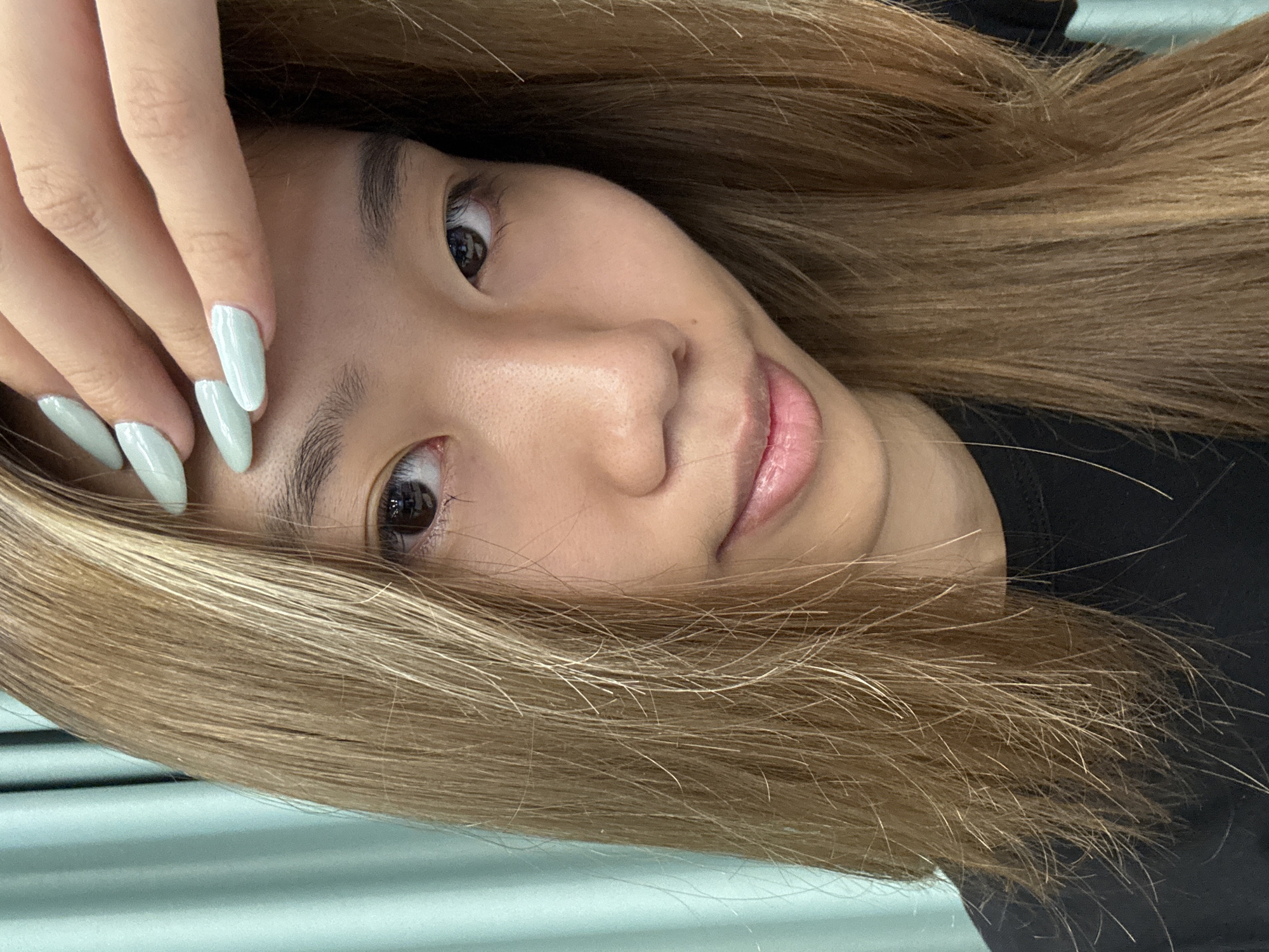 A person with light brown hair is seen with their face facing the camera. The person's left hand is raised to the upper left of the frame. The fingernails are painted a light mint green. The background has indistinct horizontal lines, and the person is wearing a black shirt. There is no information present to indicate the location is Nan'an District, China.