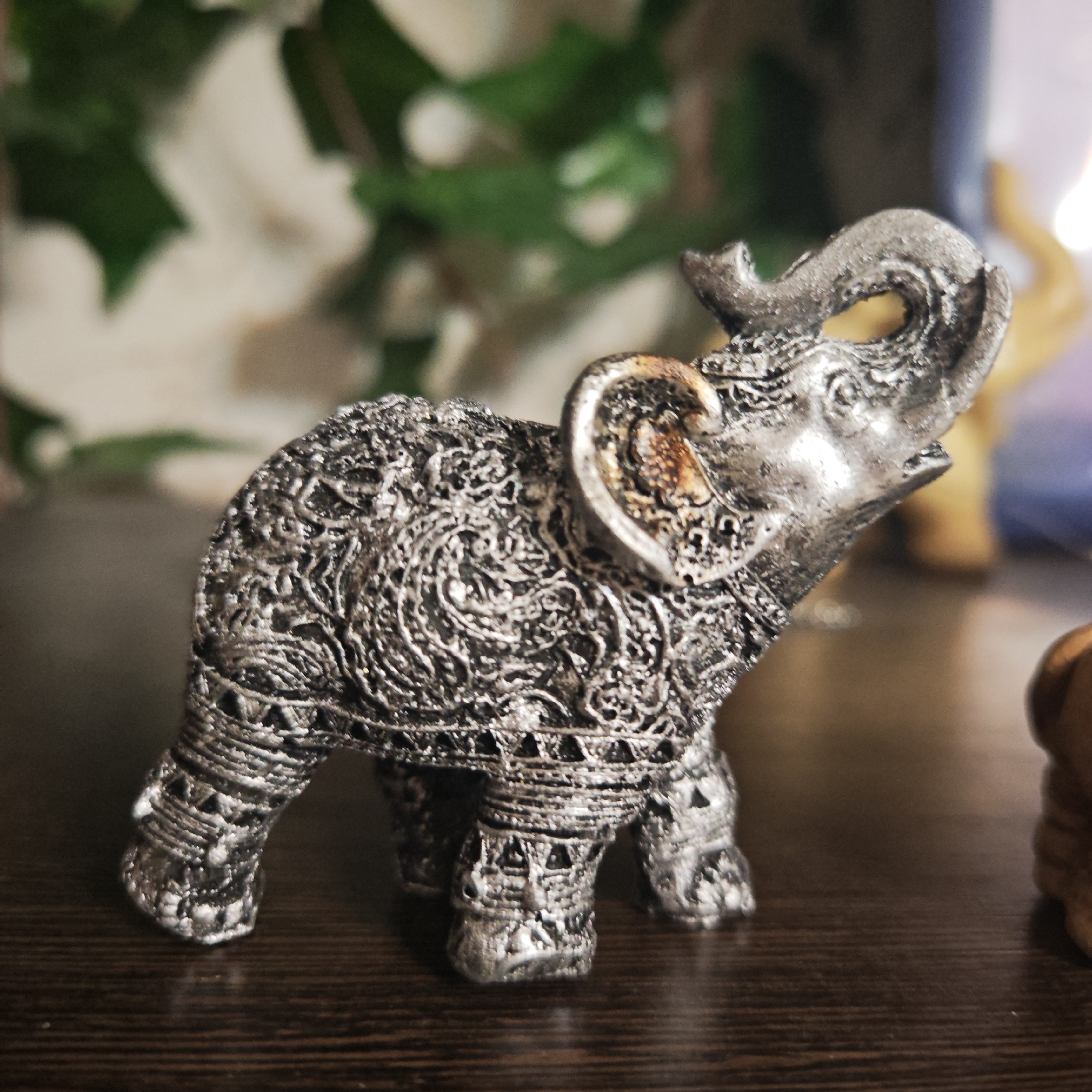 A close-up shot presents a small, intricately patterned, silver-colored elephant figurine. The elephant is positioned on a dark brown wooden surface. The foreground is in sharp focus, highlighting the ornate details of the figurine. The background is blurred, with green foliage and a hint of other figurines in the distance. There is no verifiable evidence the location is Rostov, Russia.
