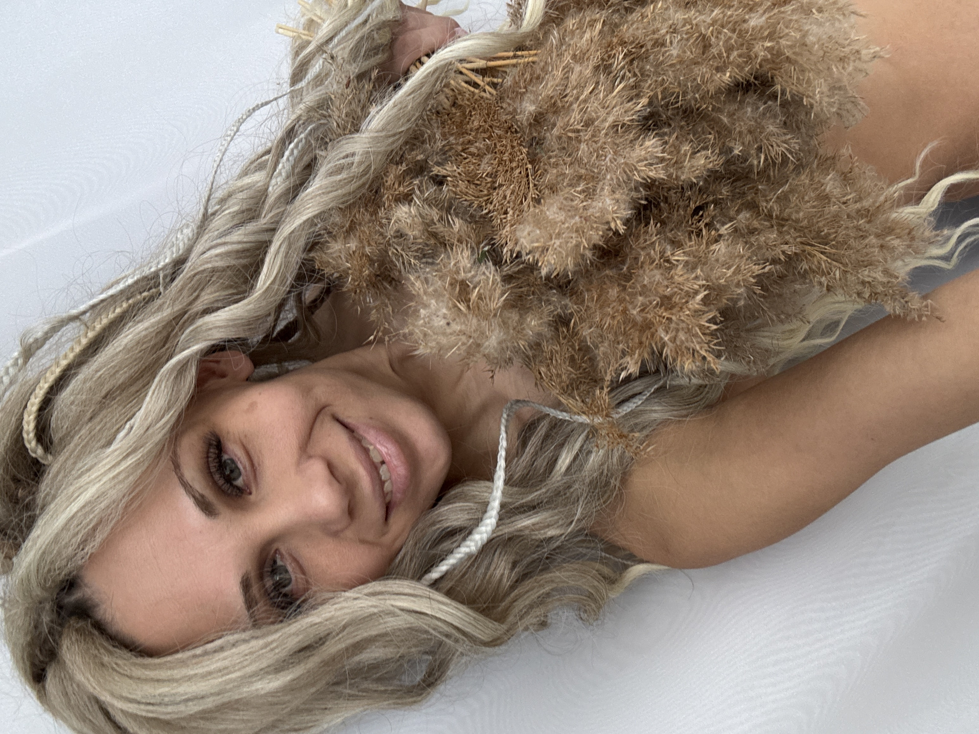 A fair-skinned woman with blonde and white braided hair lies on a white surface, likely a bed or floor, holding a bundle of dried pampas grass. Her right arm extends toward the viewer. She is smiling. This image was taken in an unknown place, but the instructions suggest Rusanovo, Russia. The specific location is unknown.