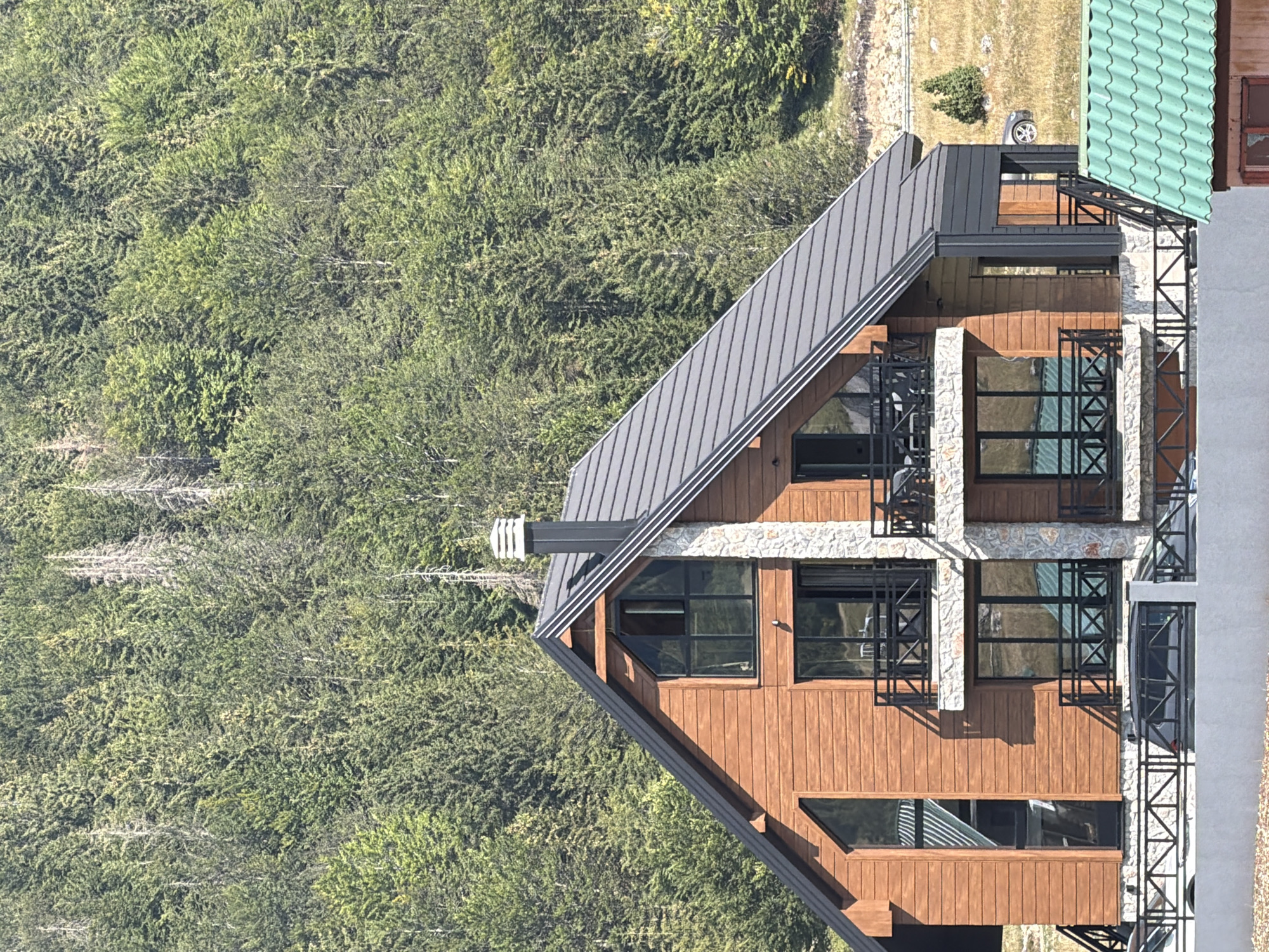 A modern wooden house with a stone facade is situated at the base of a densely forested mountainside in Virak, Montenegro. The house features a dark gray, gabled roof, multiple balconies with black railings, and large windows that reflect the surrounding greenery. A prominent stone chimney rises from the roof. The foreground shows a white retaining wall, a black metal fence, and the roof of a smaller, green-roofed structure. A car is visible parked on a dirt road to the right of the house. The environment is characterized by a steep incline covered in a variety of evergreen trees, with patches of dry grass and rocky terrain visible in the distance. The scene is bathed in natural sunlight, casting shadows on the house and mountain.