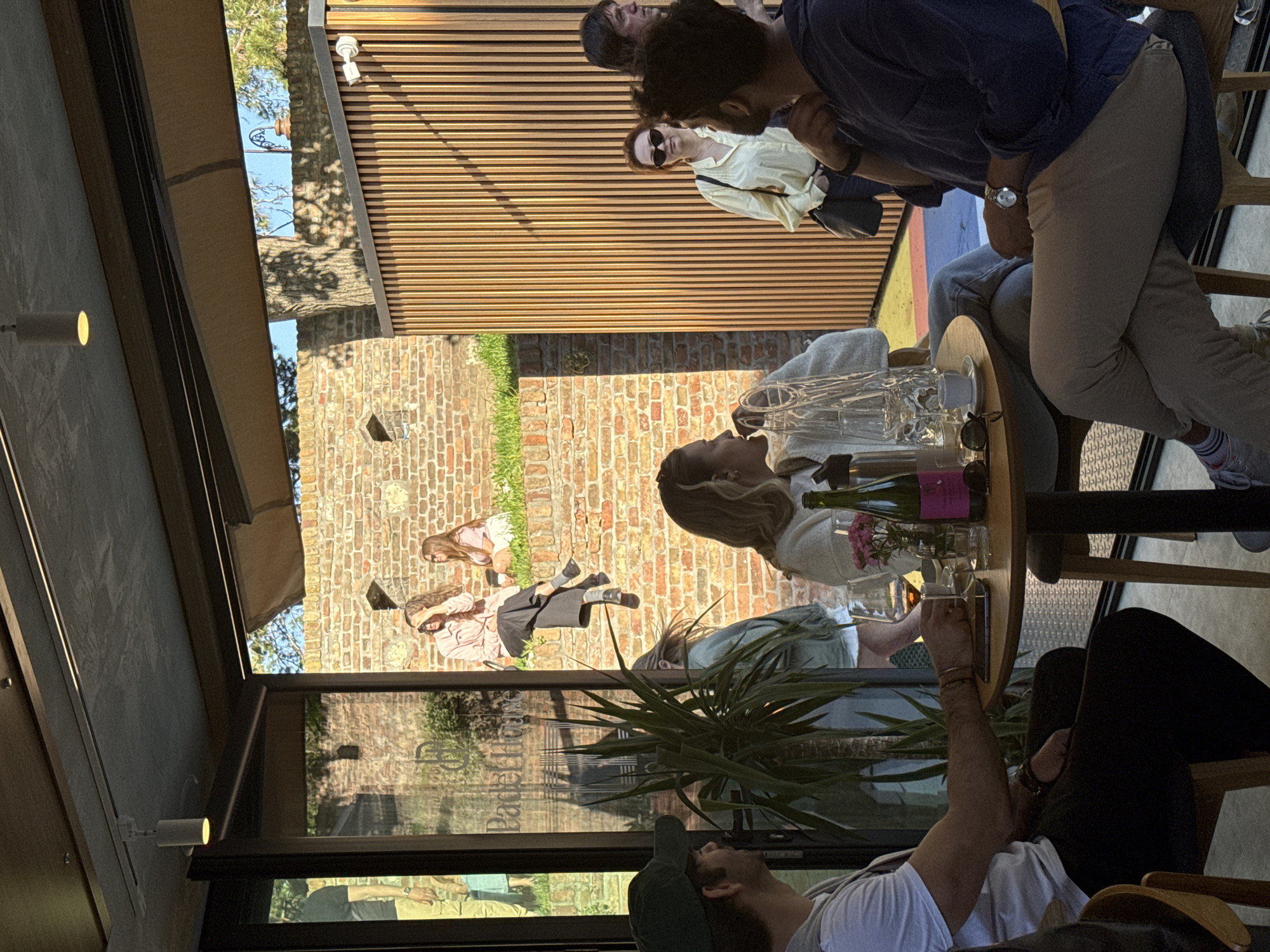 The scene appears to be set at the Bašta Bar in Belgrade, Serbia, as evidenced by a sign reflected in the glass. The setting suggests an outdoor or partially enclosed space, possibly a terrace or patio, with a view of a brick wall and some greenery. A group of people is present. Four people are sitting at a round table with drinks and glassware. A man is sitting on a chair, a bottle of wine sits on the table, next to glasses. The environment includes both indoor architectural elements and outdoor natural features. Sunlight casts shadows, adding to the ambiance of the scene.