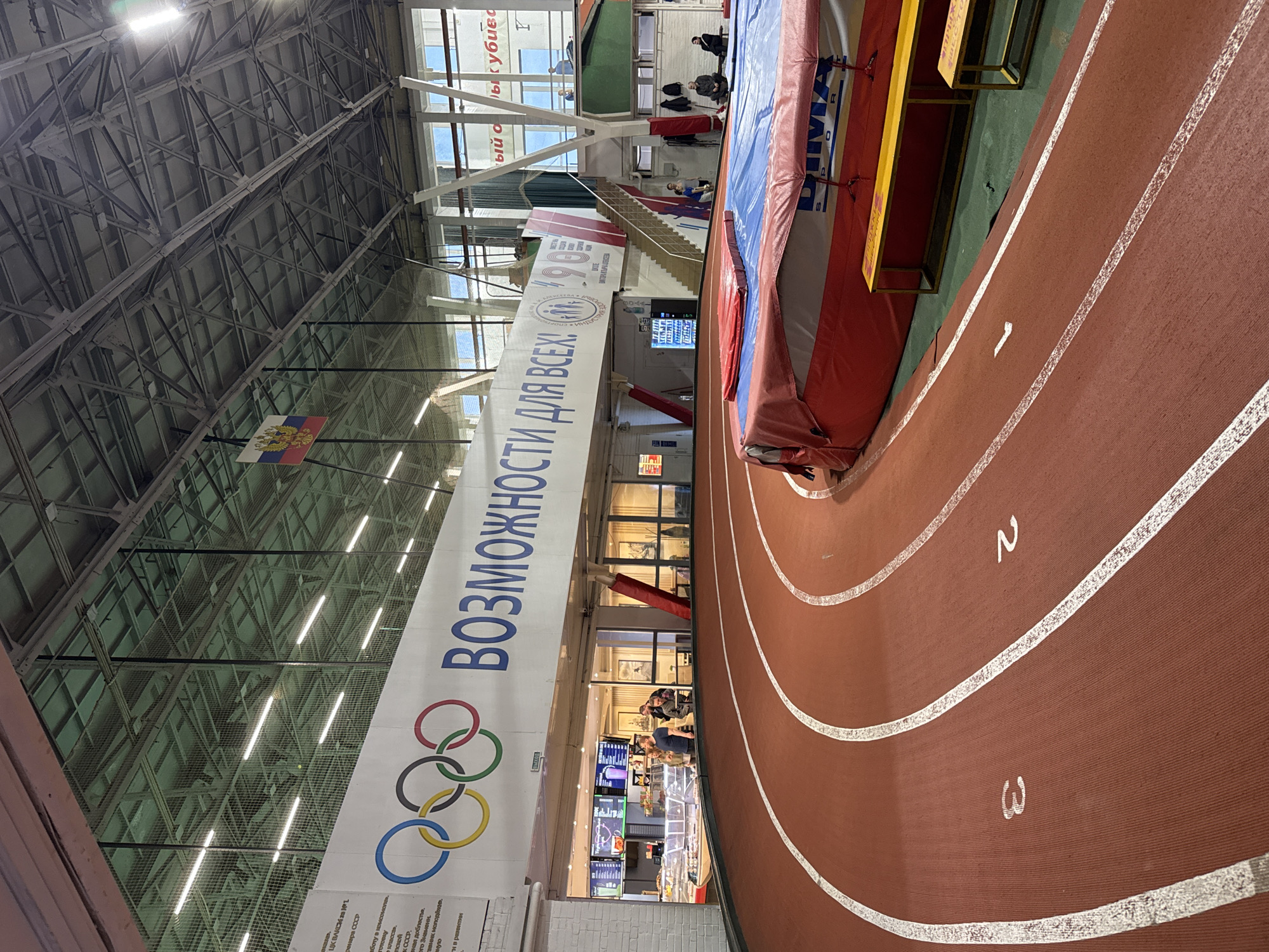 The image shows the interior of what appears to be an indoor athletics facility or sports complex. A red running track with white lane markers (numbered 1, 2, and 3) is visible. A banner hangs with text in Russian and the Olympic rings. The text translates to "Opportunities for All." Above the banner is a flag that has the emblem of Russia. Behind the banner, there is a food court or cafe visible. The ceiling is high and features a metal framework. There is also a high jump mat on the right-hand side, and the area has some natural light. Based on the information in the OCR text, this location is in Murino, Russia.