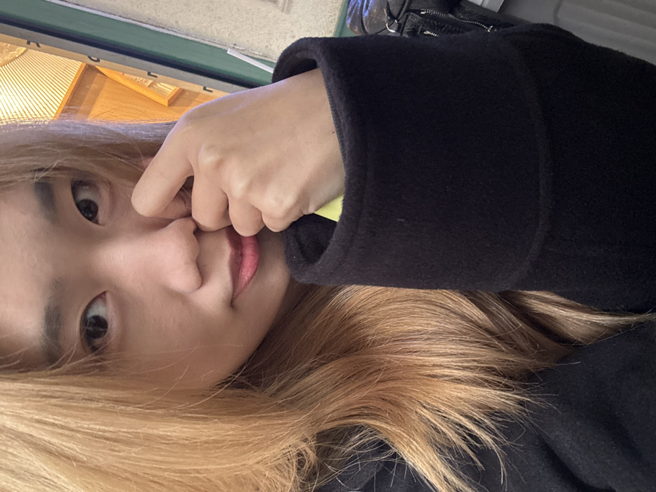 The image depicts a close-up portrait of one individual, an East Asian female, with light blonde-dyed hair and a fair complexion. Her dark eyes are directed forward, and her lips are slightly parted, exhibiting a subtle smile and bearing red or pink lipstick. She is dressed in a dark-colored, heavy-gauge fabric garment, consistent with a coat or jacket, featuring a prominent cuff on the right arm. Her right hand is raised, with the index finger and thumb lightly touching the right side of her nose. A small, bright yellow object is partially visible inside the cuff of her right sleeve. The background is largely out of focus but reveals elements indicative of an indoor setting, including a vertical green strip and a light-colored, possibly wooden or textured, slatted partition. A black object with a visible zipper, potentially a bag, is partially visible on the right side of the frame. This scene is located in Yuzhong District, China.