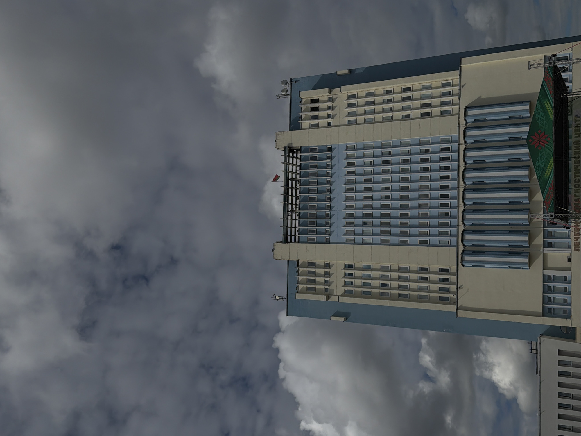 The image shows a tall, modern building with a distinctive architectural style, likely located in the city of Mahilyow, Belarus. The building's facade is a combination of light cream and pale blue vertical sections, punctuated by numerous small rectangular windows. The upper part of the building features a more elaborate design with a row of protruding vertical elements and what appear to be communication antennas. A small flag, possibly a national or regional emblem, is visible flying from the rooftop. Below the main structure, a stage with a green canopy decorated with a red and green emblem is set up in front of a lower section of the building. The canopy has text on it, part of which reads "ДИАГНОСТИЧЕСКИЙ ЦЕНТР", indicating it is a diagnostic center. The sky is filled with dramatic, dark clouds, suggesting overcast weather. The overall impression is one of a functional, possibly institutional building set against a moody sky.
