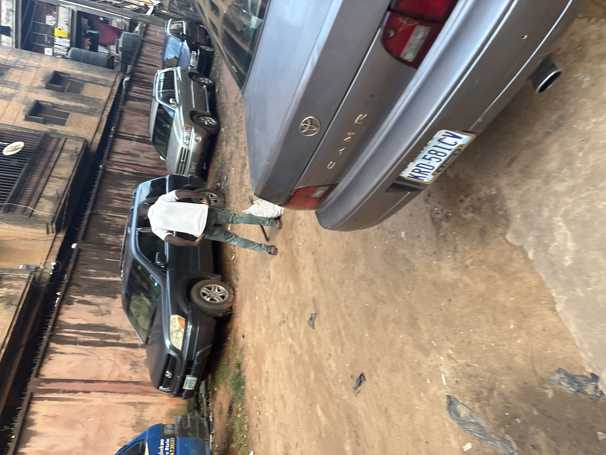 The image captures a street scene in Onitsha, Nigeria, characterized by a dirt ground and several parked vehicles. In the foreground, a gray Toyota Camry is partially visible, with its rear end facing the camera. The license plate reads "KRD 581CV". To the left of the Camry, a dark-colored SUV is parked, and behind it, a silver SUV. Further to the left, a blue van is also parked. A man dressed in a white shirt and green pants is standing near the dark SUV, holding a white bag. He appears to be engaged in an activity on the ground, possibly cleaning or collecting debris. The environment suggests an urban setting with buildings visible in the background, including balconies with laundry hanging. The overall scene depicts a typical street in a Nigerian city, with elements of daily life and the presence of various vehicles.