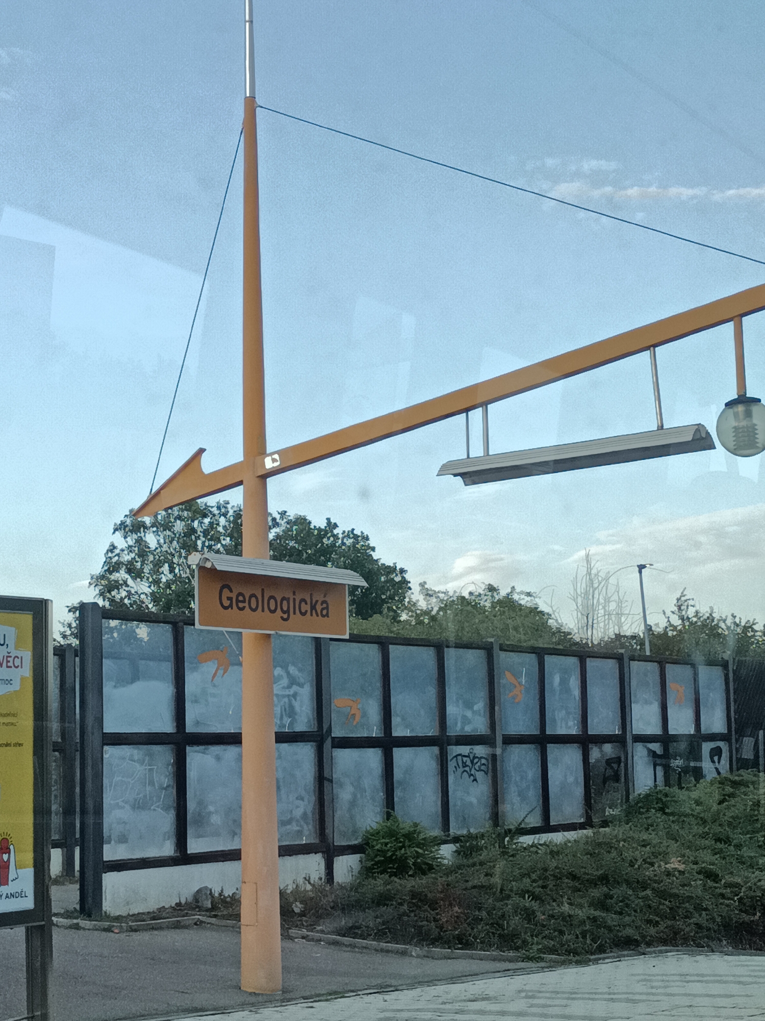 The image depicts a sign in the city of Prague, Czechia, that reads "Geologická". This sign is mounted on an orange pole, which is part of a larger structure with orange beams and what appears to be a light fixture and a lamp attached. The sign itself is brown with white lettering. Behind the sign and pole, there is a glass sound barrier with dark frames. Orange bird-like silhouettes are visible on the glass panels of the sound barrier. Graffiti is present on some of the glass panels. Green foliage and trees are seen behind the sound barrier. The sky above is light blue with some clouds. In the lower left corner, a portion of a yellow sign is visible with text and an illustration of a figure. The scene appears to be outdoors, possibly near a street or public transport stop.