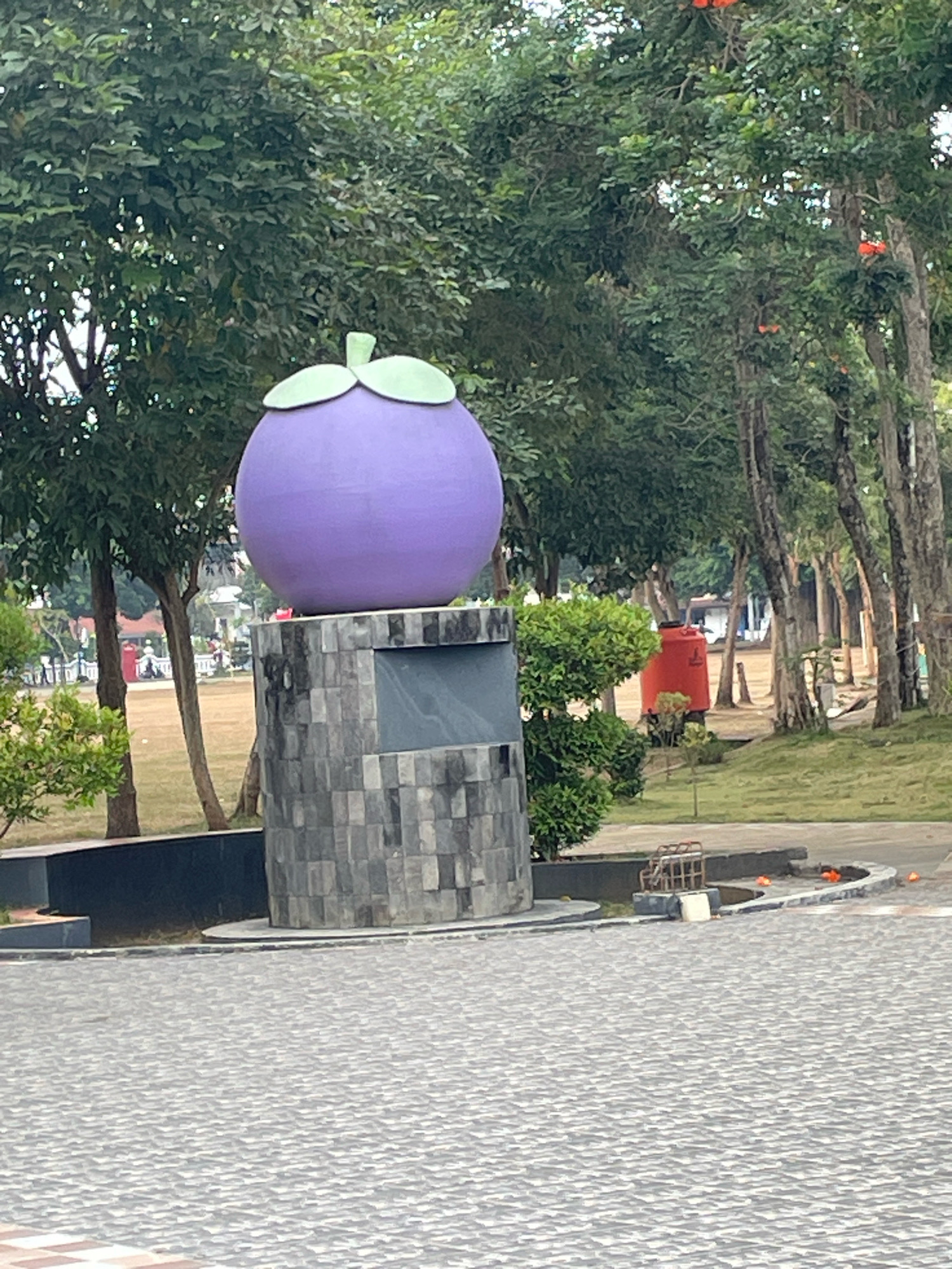 A large, purple, spherical statue with green leaves on top, resembling a mangosteen, stands on a stone pedestal in a park in Purworejo, Indonesia. The pedestal is made of grey, rectangular stones arranged in a pattern. In the background, there are trees with green foliage and some bare branches. Orange-colored objects, possibly cones or flowers, are scattered on the ground near a metal structure with a white object. A large, red water tank is visible behind a green bush. The ground in the foreground is paved with grey interlocking stones. In the distance, there are buildings and a few people walking. The environment appears to be a public park or recreational area.
