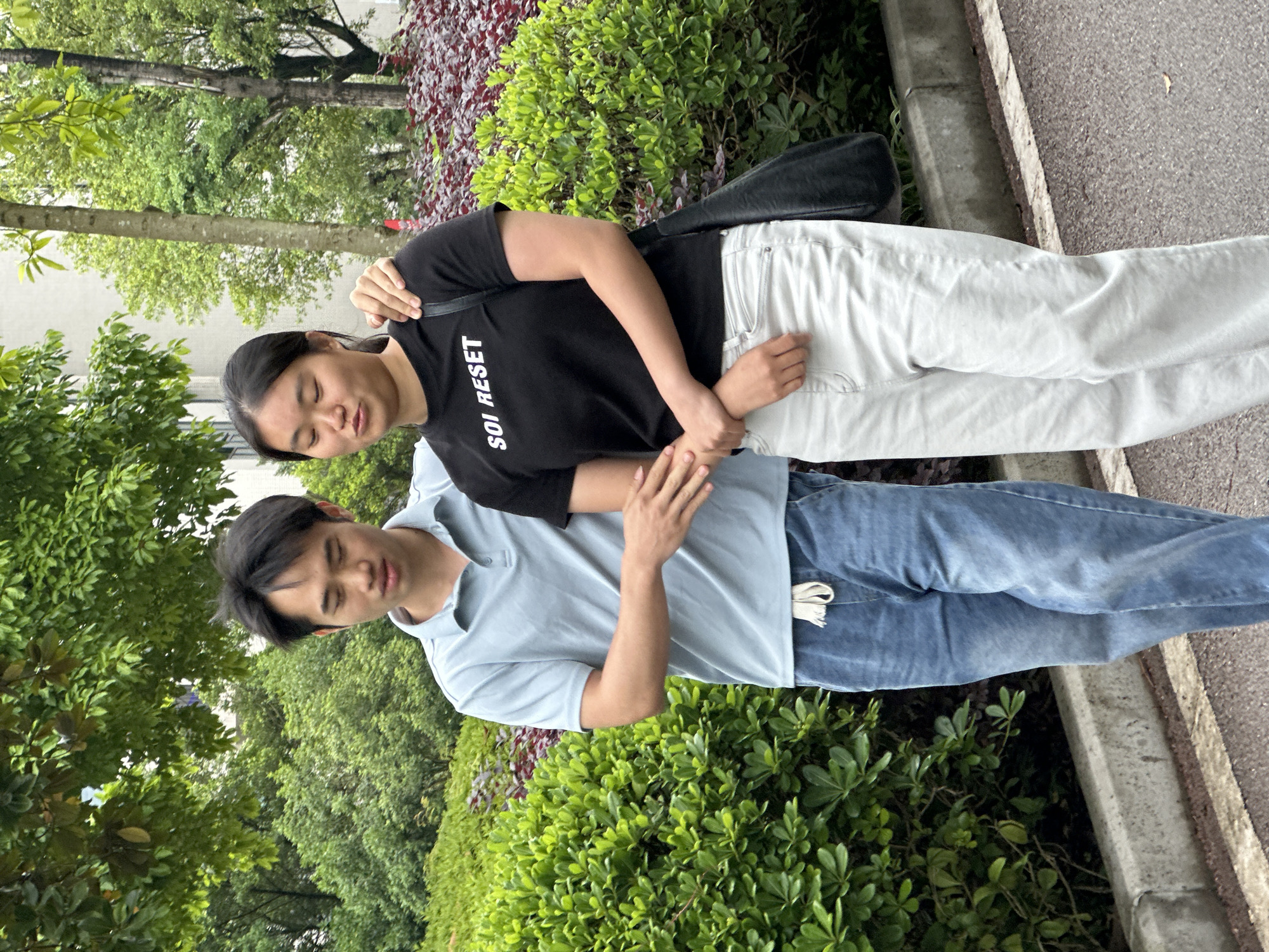 A young Asian man and woman are standing next to a sidewalk in Jiangjin, China. The woman has her arm around the man's shoulder and is wearing a black t-shirt with the words "SOI RESET" and white jeans. The man is wearing a light blue polo shirt and blue jeans. They are standing next to a green bush. A sidewalk is located to their right. Trees are visible in the background.