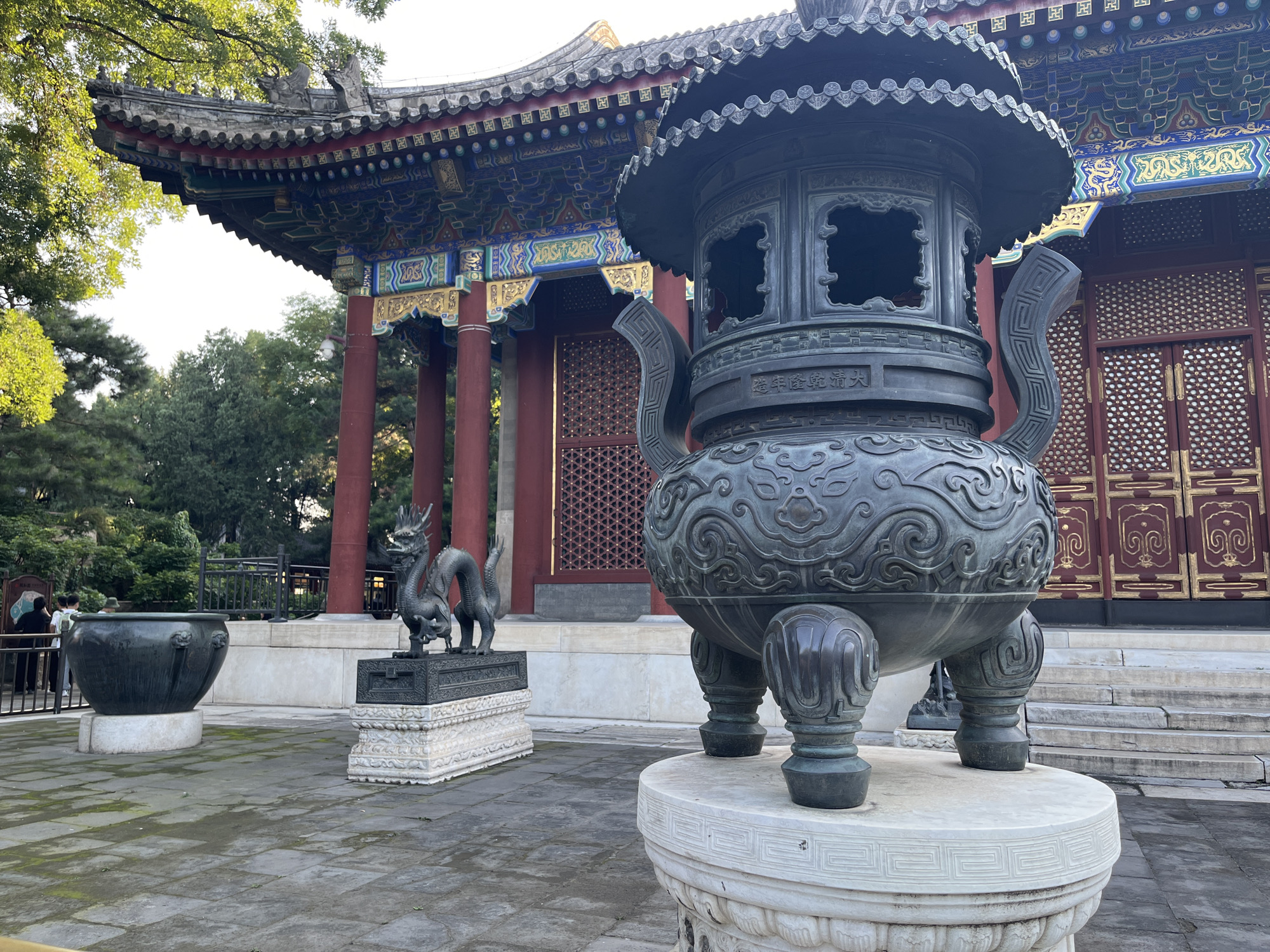 An outdoor, eye-level, medium shot captures a courtyard in front of a traditional Chinese building. The building features a red wooden facade with ornate gold and dark red latticework, topped by a multi-tiered roof with grey tiles and vibrant blue, green, and red painted eaves. Red columns support the eaves. In the foreground, a large, dark bronze incense burner with intricate carvings rests on a white pedestal. To the left, a smaller, dark bronze basin sits on another white base. Behind the basin, a dark bronze statue of a dragon stands on a decorative stone platform. Further to the left, two individuals are visible near a fence. The courtyard is paved with square grey stones, some of which are covered in moss. Lush green trees and foliage surround the scene.