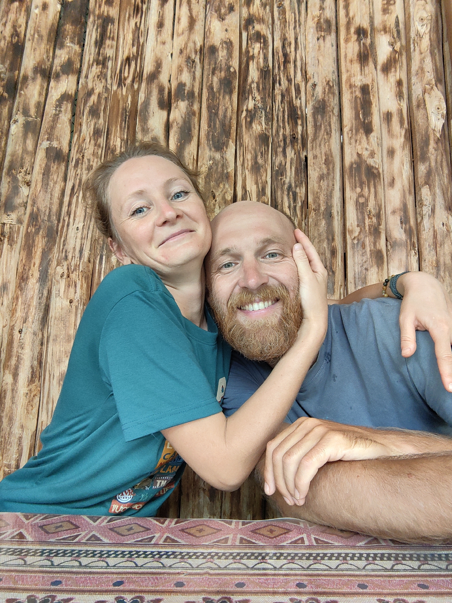 A couple poses for a close-up photo in Thành phố Hà Giang, Vietnam, against a backdrop of vertical wooden planks with dark burn marks. The woman, on the left, wears a teal t-shirt and has light hair pulled back. She has a slight smile and embraces the man. The man, on the right, is bald with a red beard and a broad smile. He wears a blue-grey t-shirt and has a beaded bracelet on his wrist. A patterned rug with geometric designs and warm colors lies in the foreground.